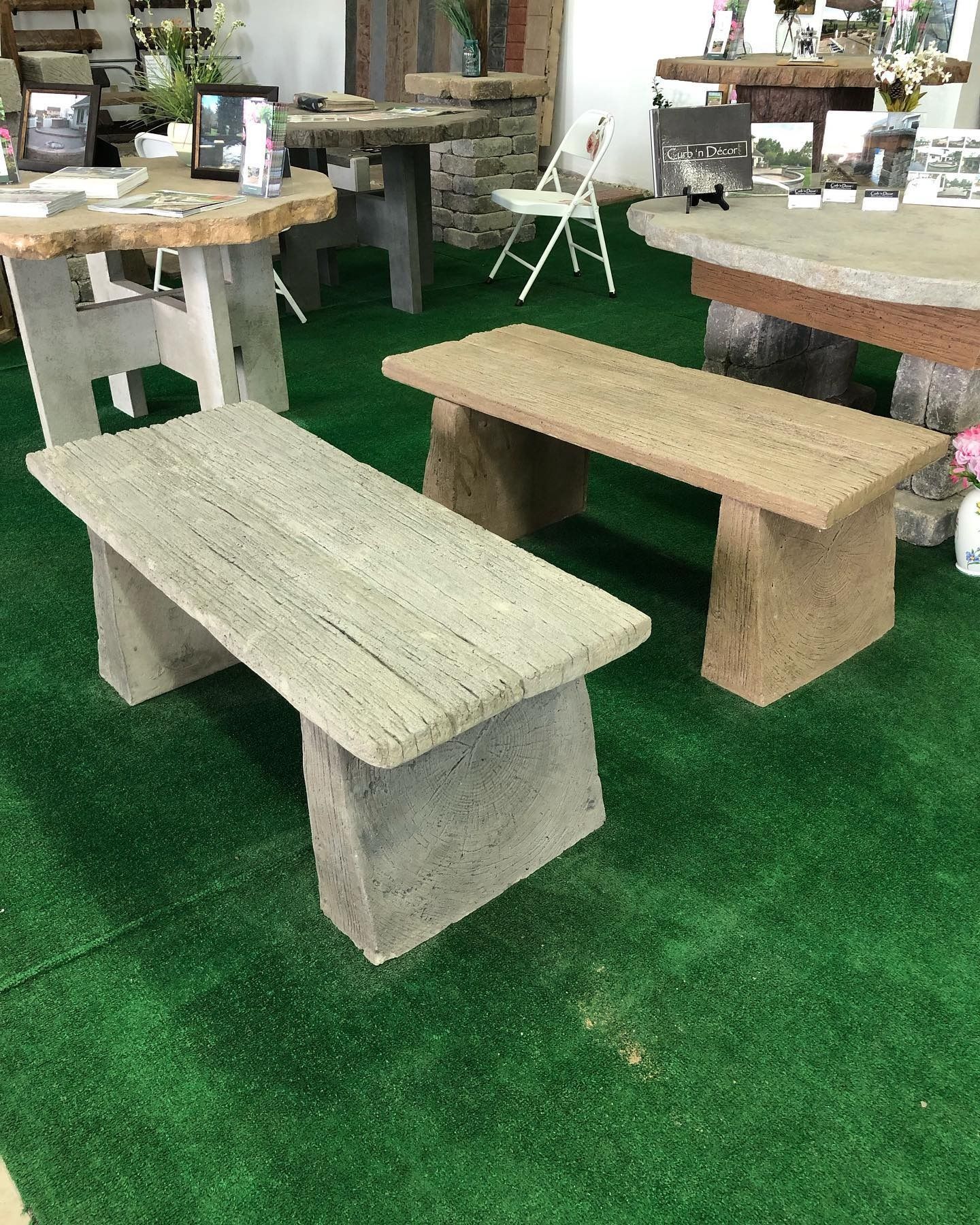 Two stone benches in gray and tan sit on green artificial turf in a showroom filled with stone patio furniture.