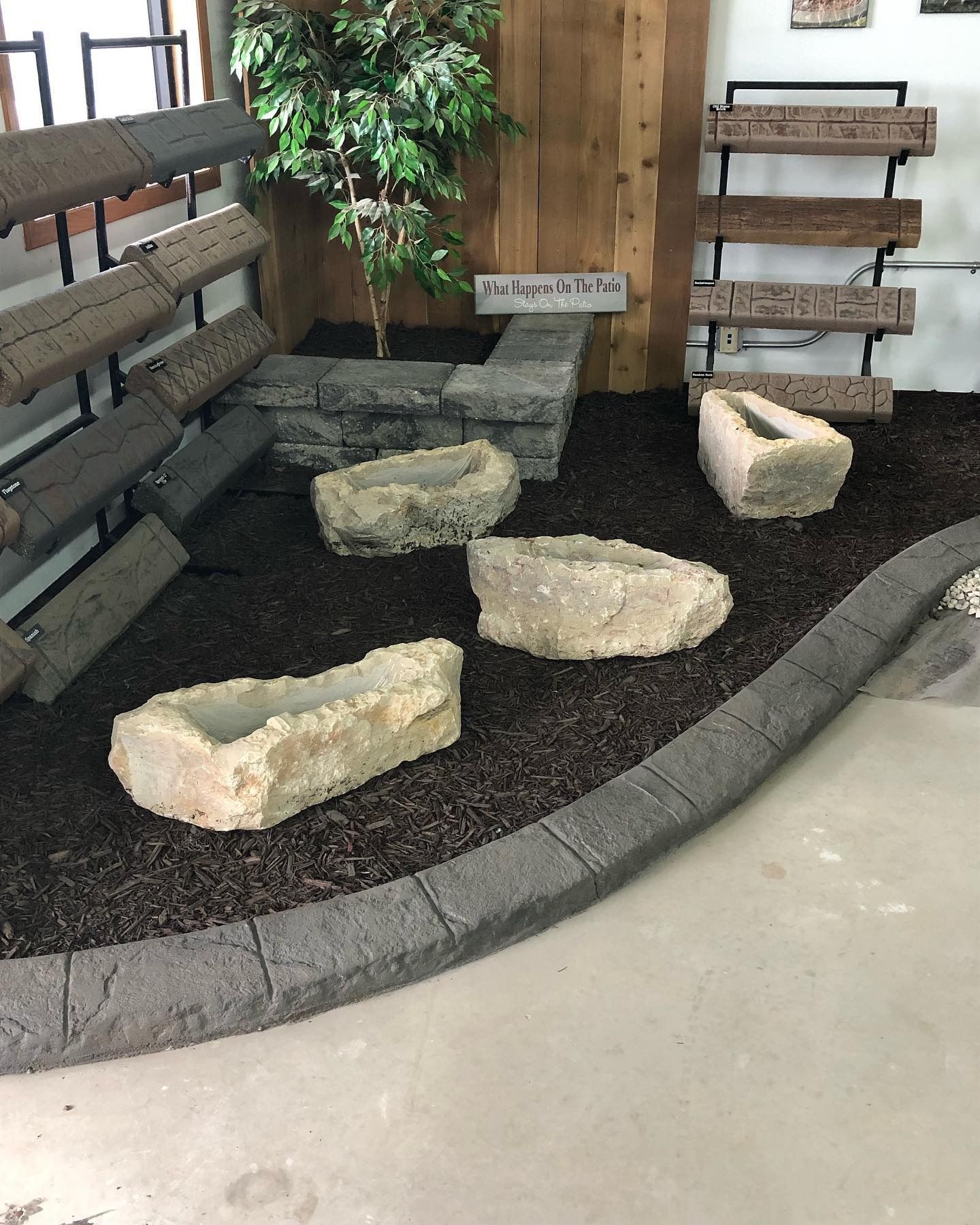 A display of stone landscape products, featuring planters on mulch and brick edging in an indoor retail setting.