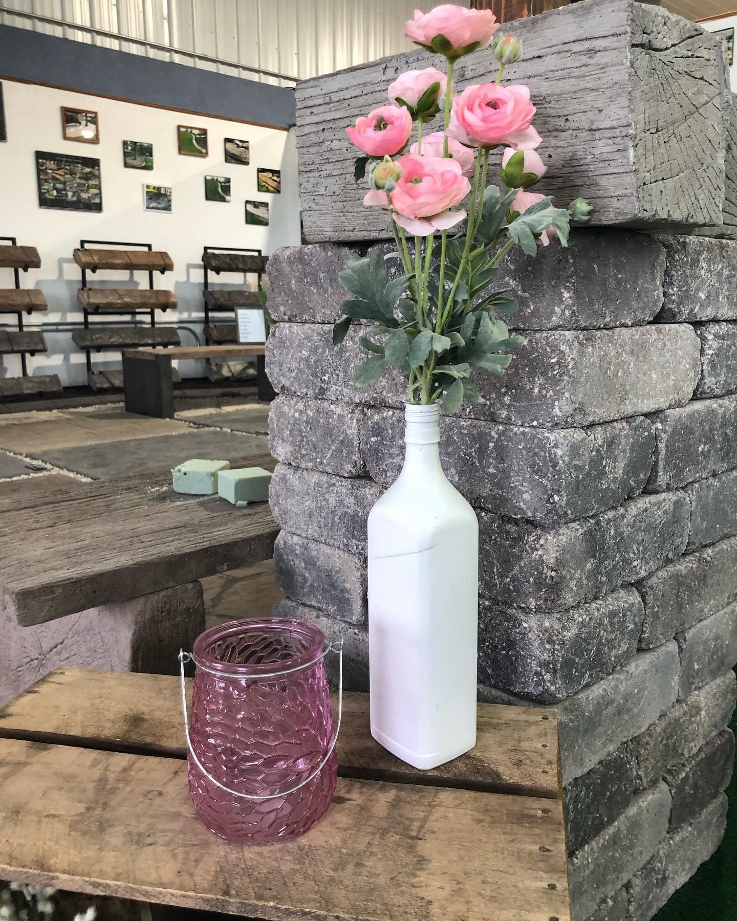 A white bottle vase with pink flowers and a pink glass lantern sit on a wooden crate against a background of stone blocks.