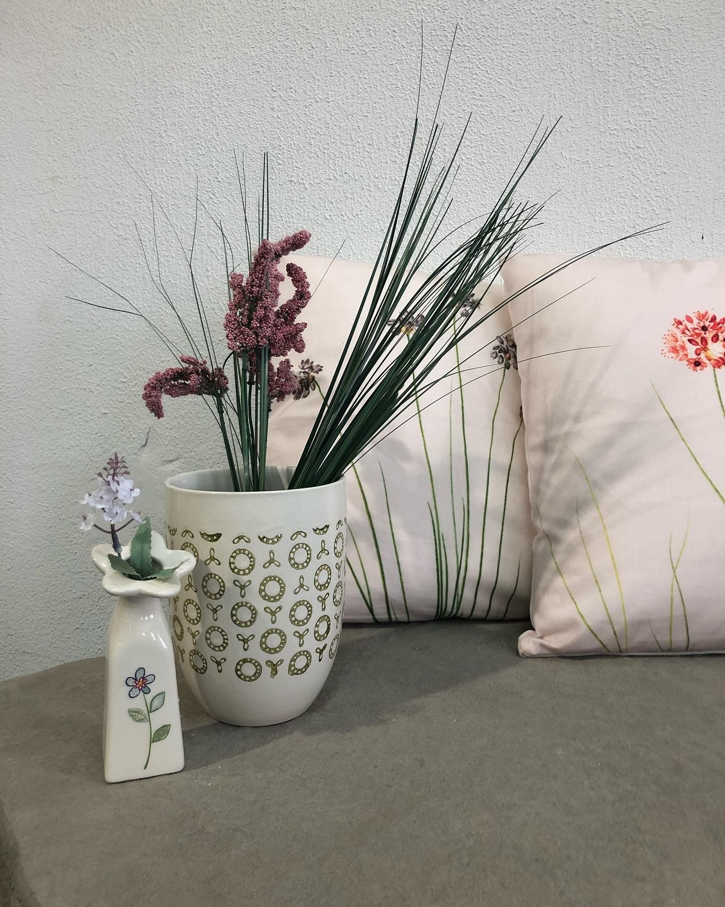 Two vases with artificial plants placed on a gray surface in front of patterned pillows against a textured wall.