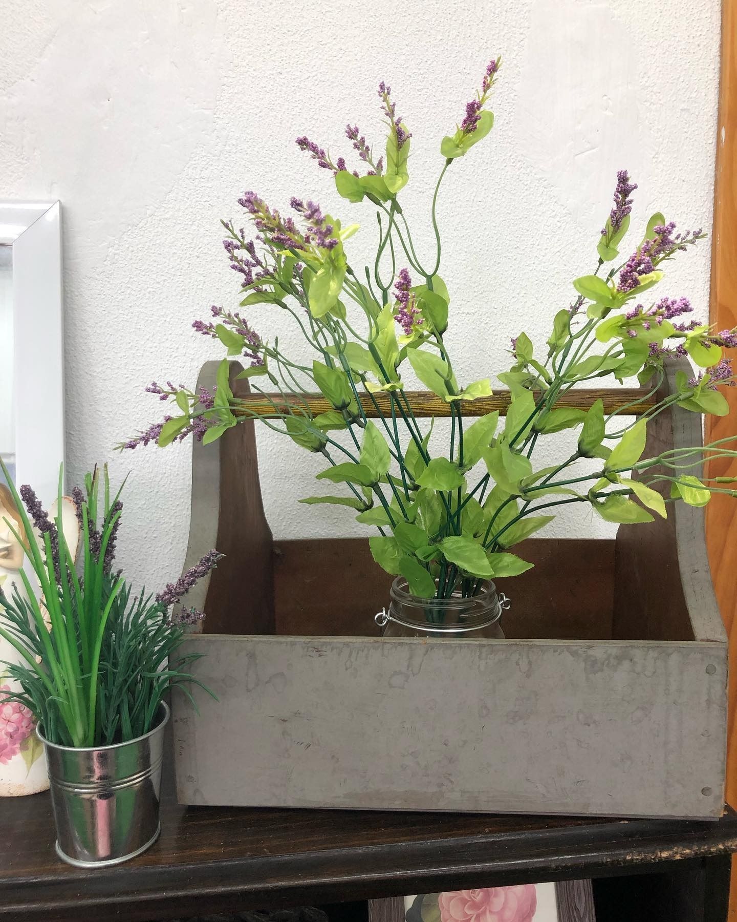 A light gray wooden caddy with floral stems inside sits next to a small metal bucket of lavender on a dark wood surface.