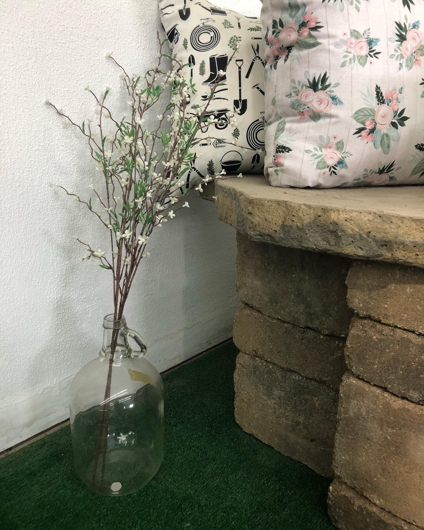 A clear glass jug holds white-flowered branches next to a stone bench with two patterned decorative pillows.