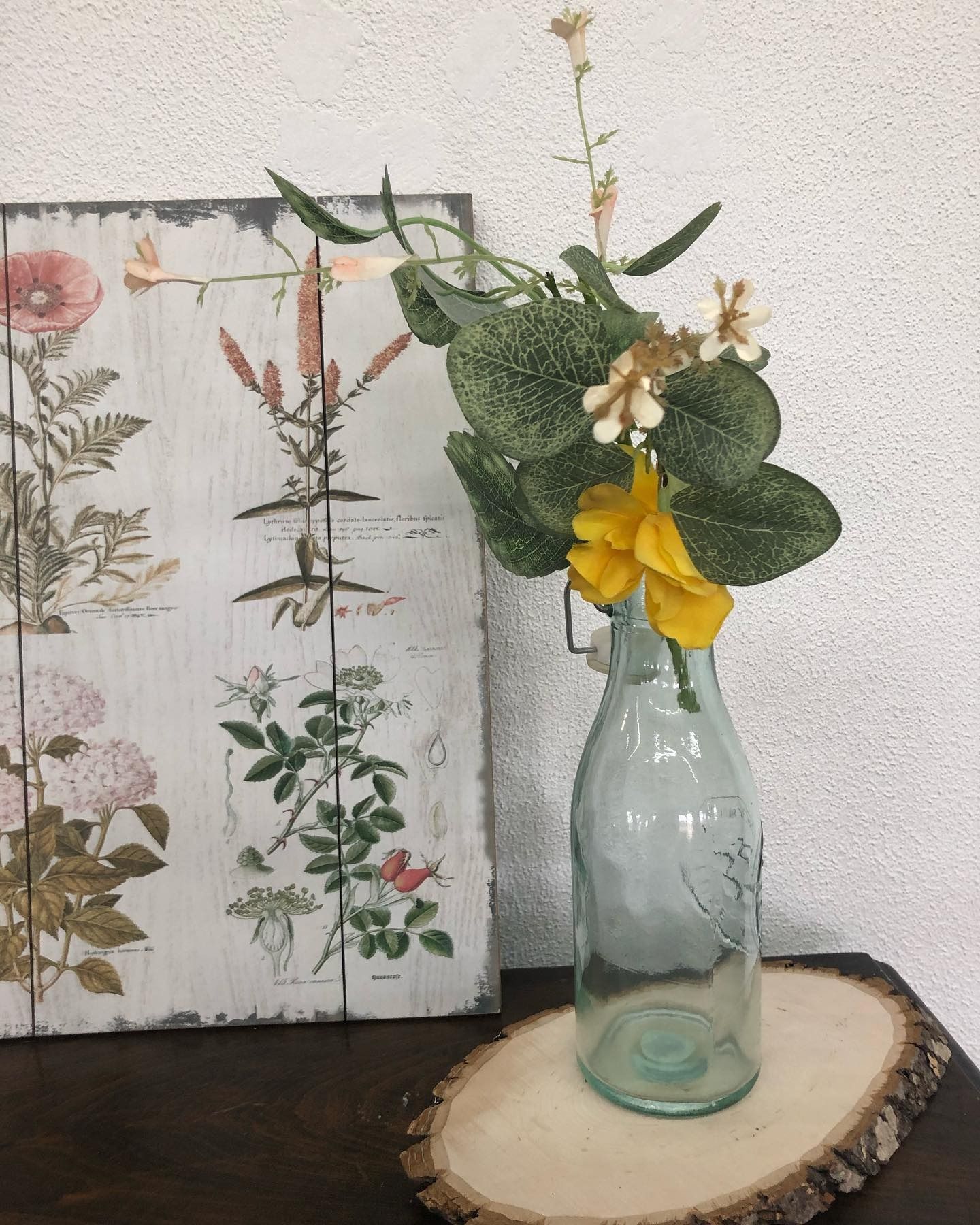 A light blue glass bottle containing faux flowers and greenery, sitting on a wood slice next to a botanical wall print.