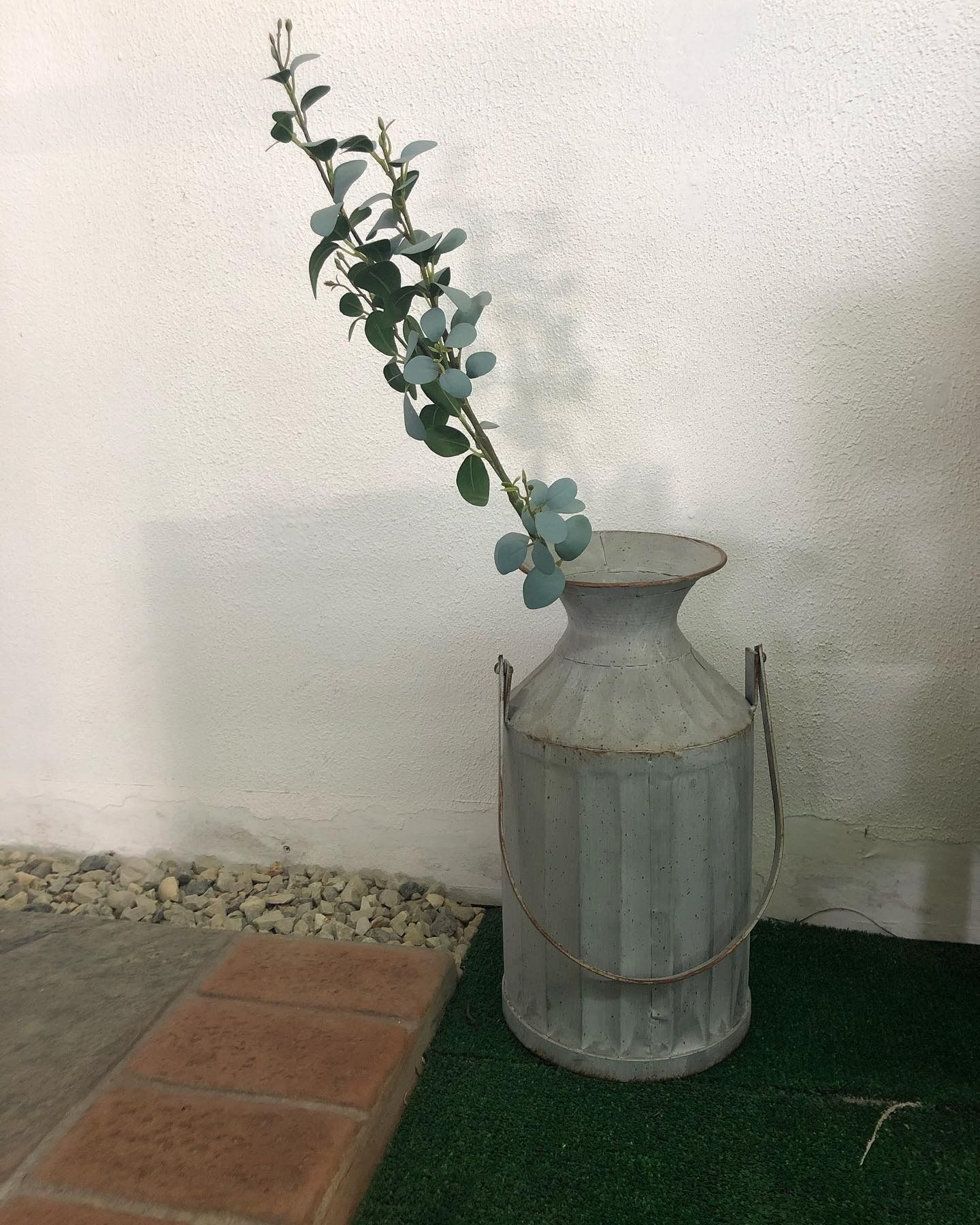 A single eucalyptus stem in a rustic, light-grey metal milk can sits on a patch of green turf against a white wall.