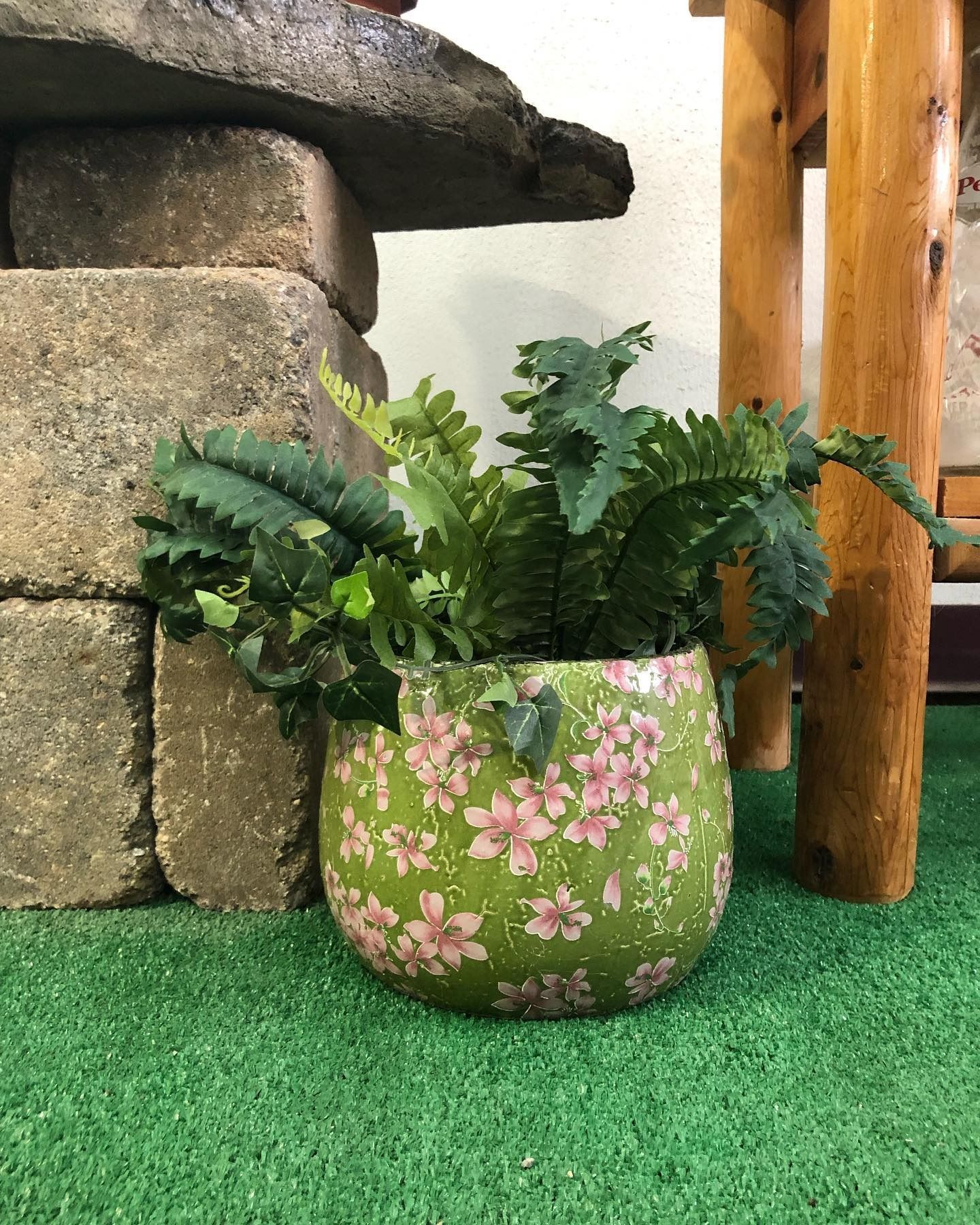 A potted fern in a green, floral-patterned ceramic pot sitting on green turf between brick blocks and a wooden post.