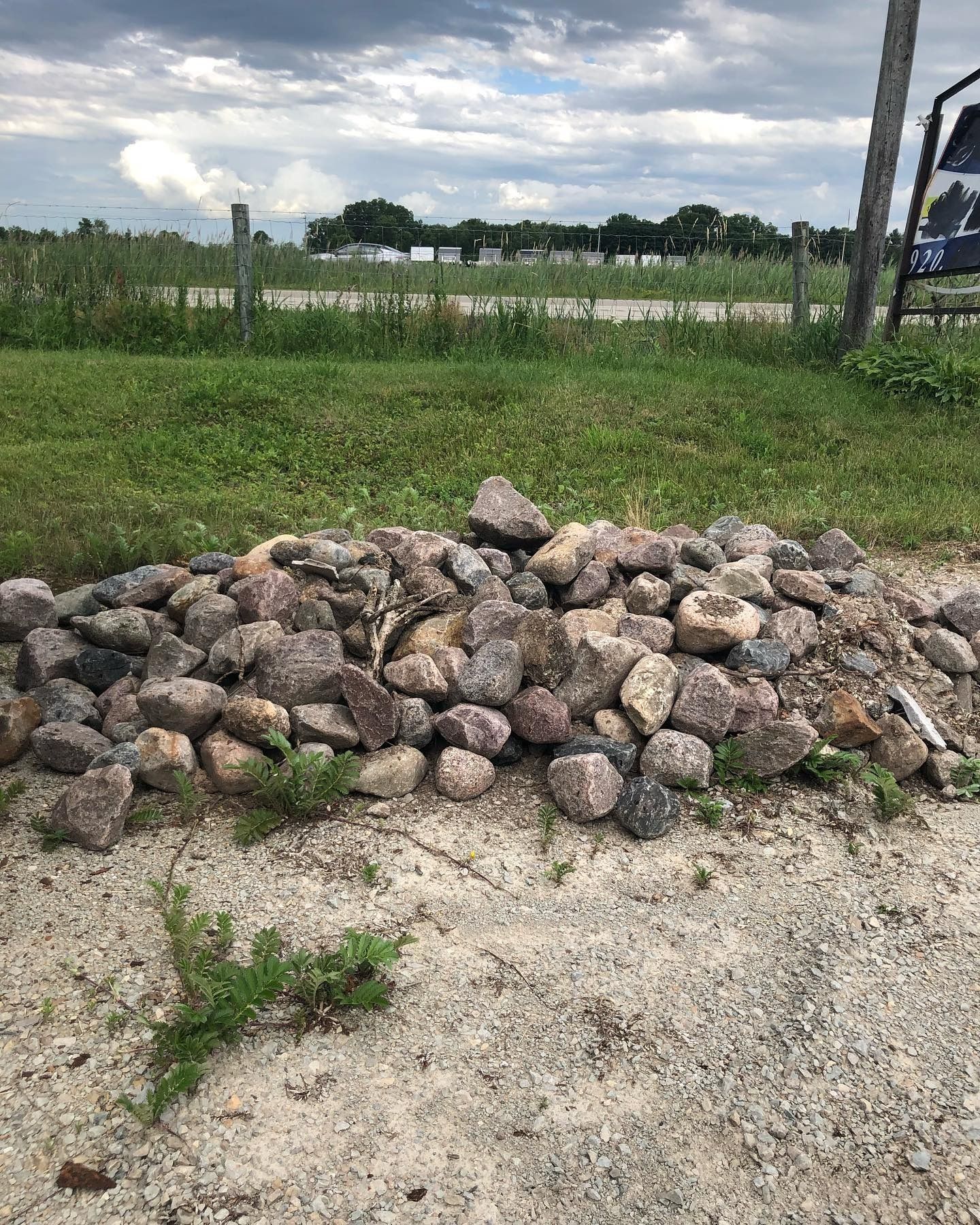 A pile of rough, multicolored rocks sits on a patch of grass next to a gravel path under a cloudy sky.