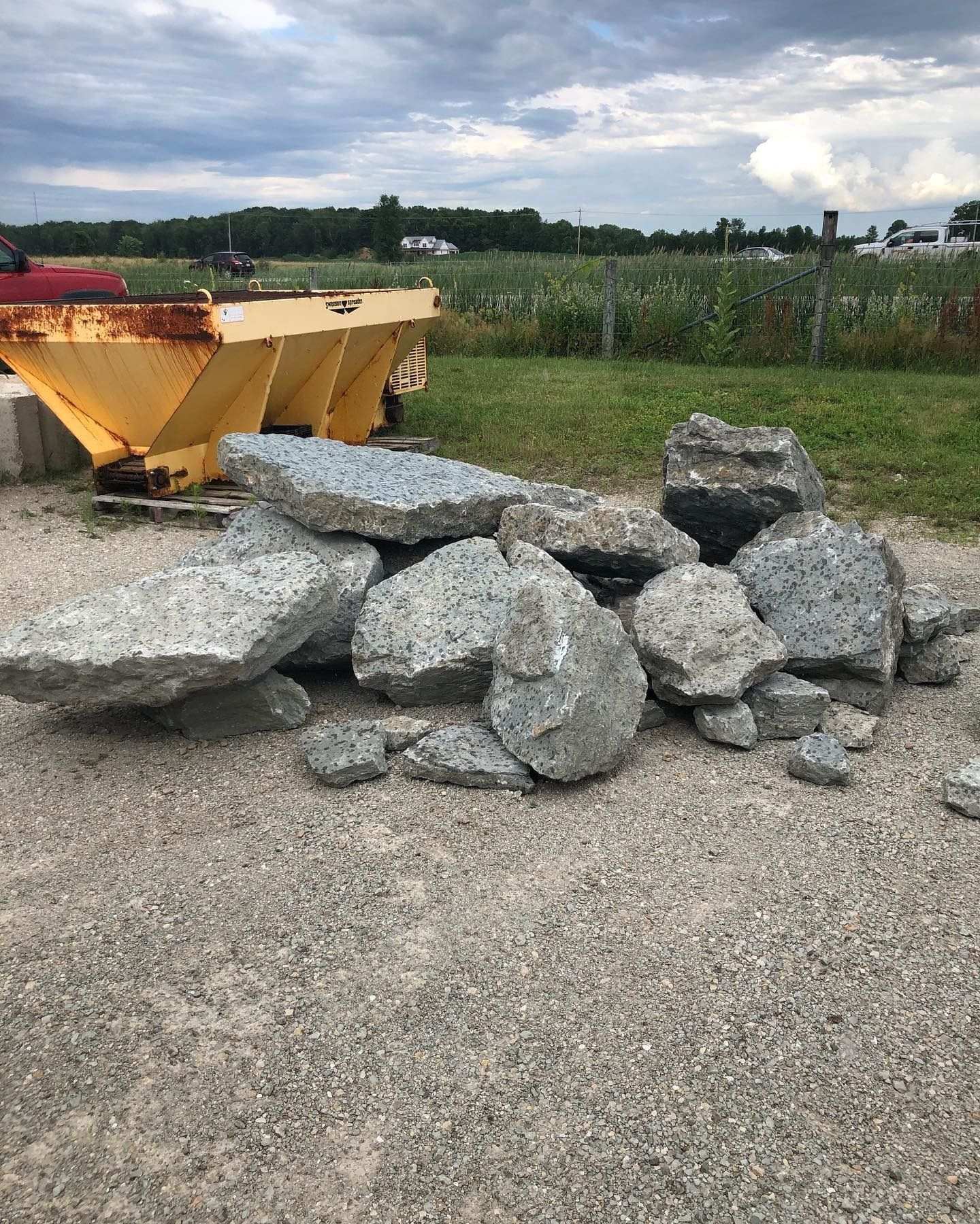 A pile of large, broken concrete chunks sits on a gravel lot near a yellow industrial hopper and a grassy field.