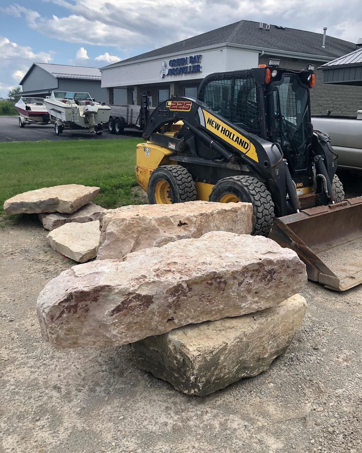 A yellow New Holland skid steer parked on a gravel lot next to several large, light-colored landscape boulders.
