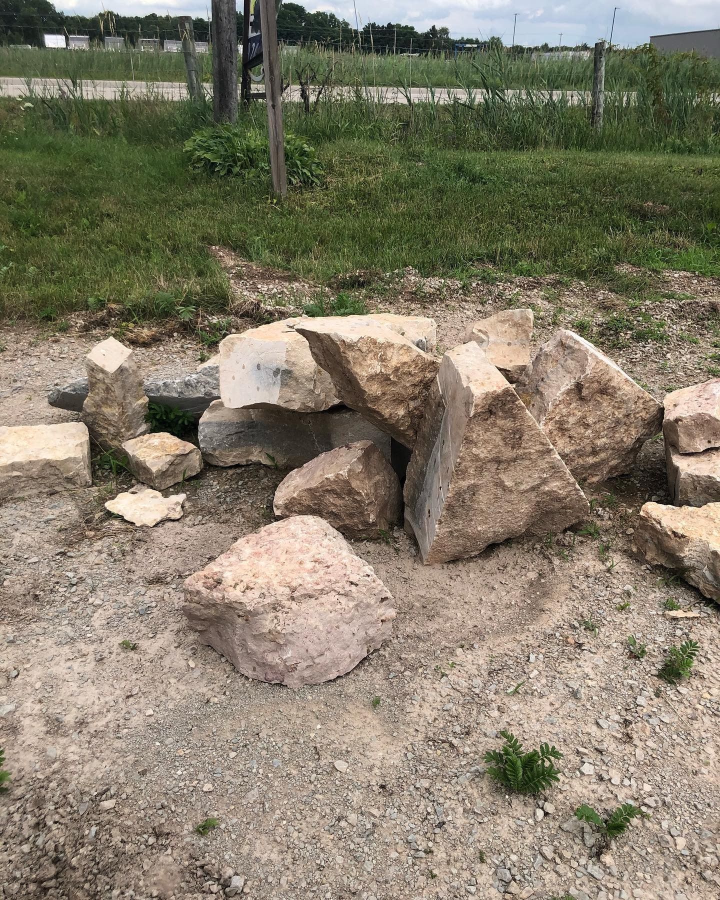 A small pile of rough-cut, light-tan limestone rocks sits on a patch of gravel next to a grassy field.