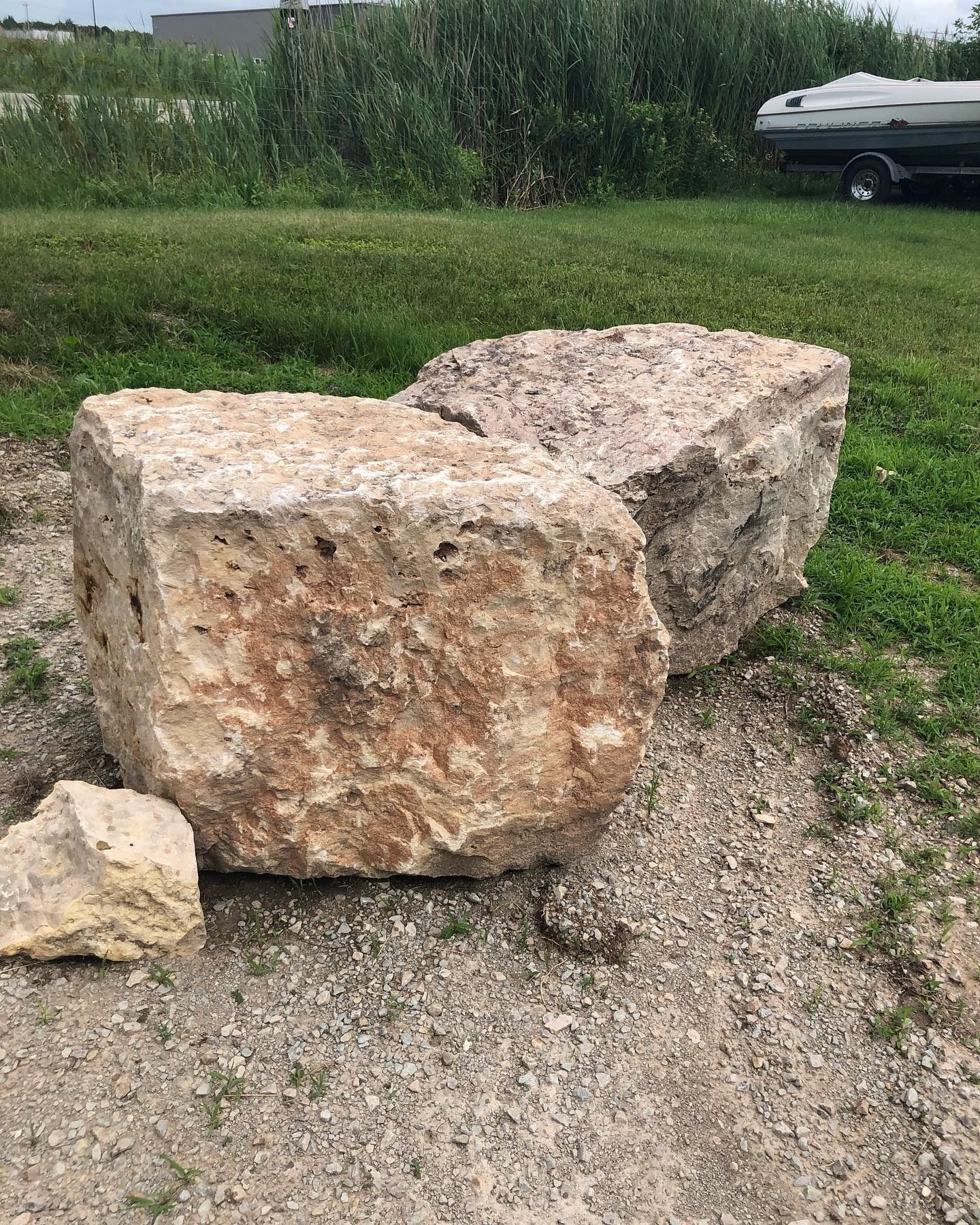 Two large, light-brown, textured limestone boulders sit on a gravel ground with grass and a boat trailer in the background.