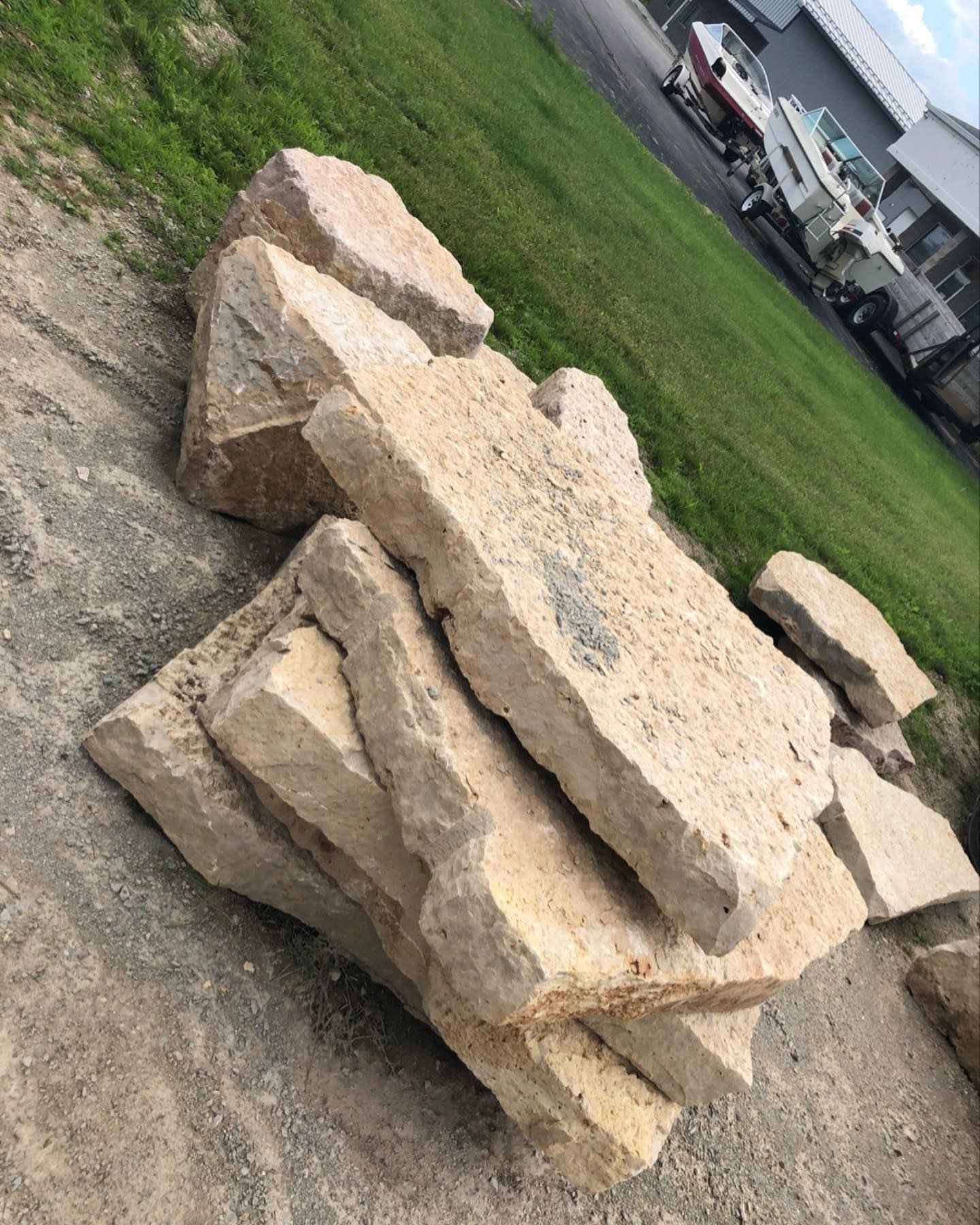 A stack of large, rough-hewn, light tan stone slabs resting on a gravel surface next to a grassy area.