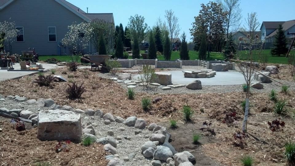 A landscaped backyard features a stone-bordered patio with a fire pit, surrounded by mulch, rock accents, and young trees.