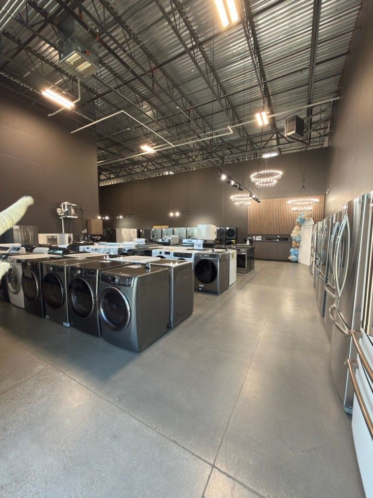 Row of front-loading washers and dryers in a laundromat with high ceilings and bright overhead lights