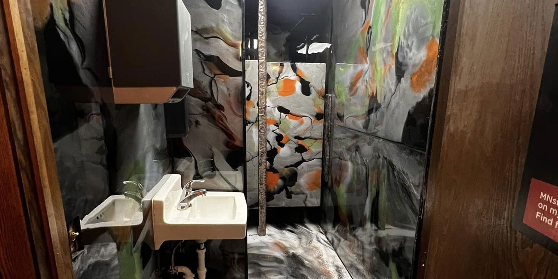 Indoor scene with a narrow mirrored walkway, white pedestal sinks, and abstract black-and-orange wall art.