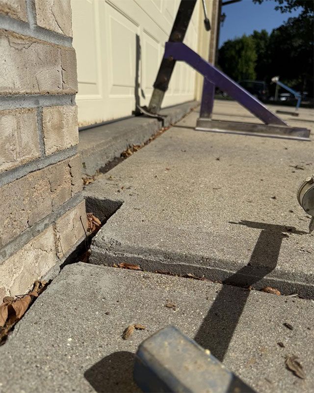 Broken concrete walkway beside a house door, with a purple strap and uneven slabs creating a trip hazard.