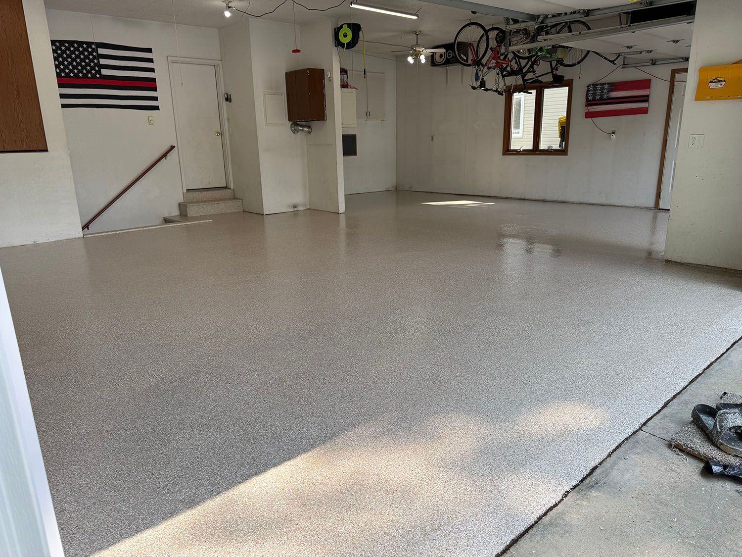 Empty garage with speckled epoxy floor, white walls, stairs, and a window letting in sunlight.