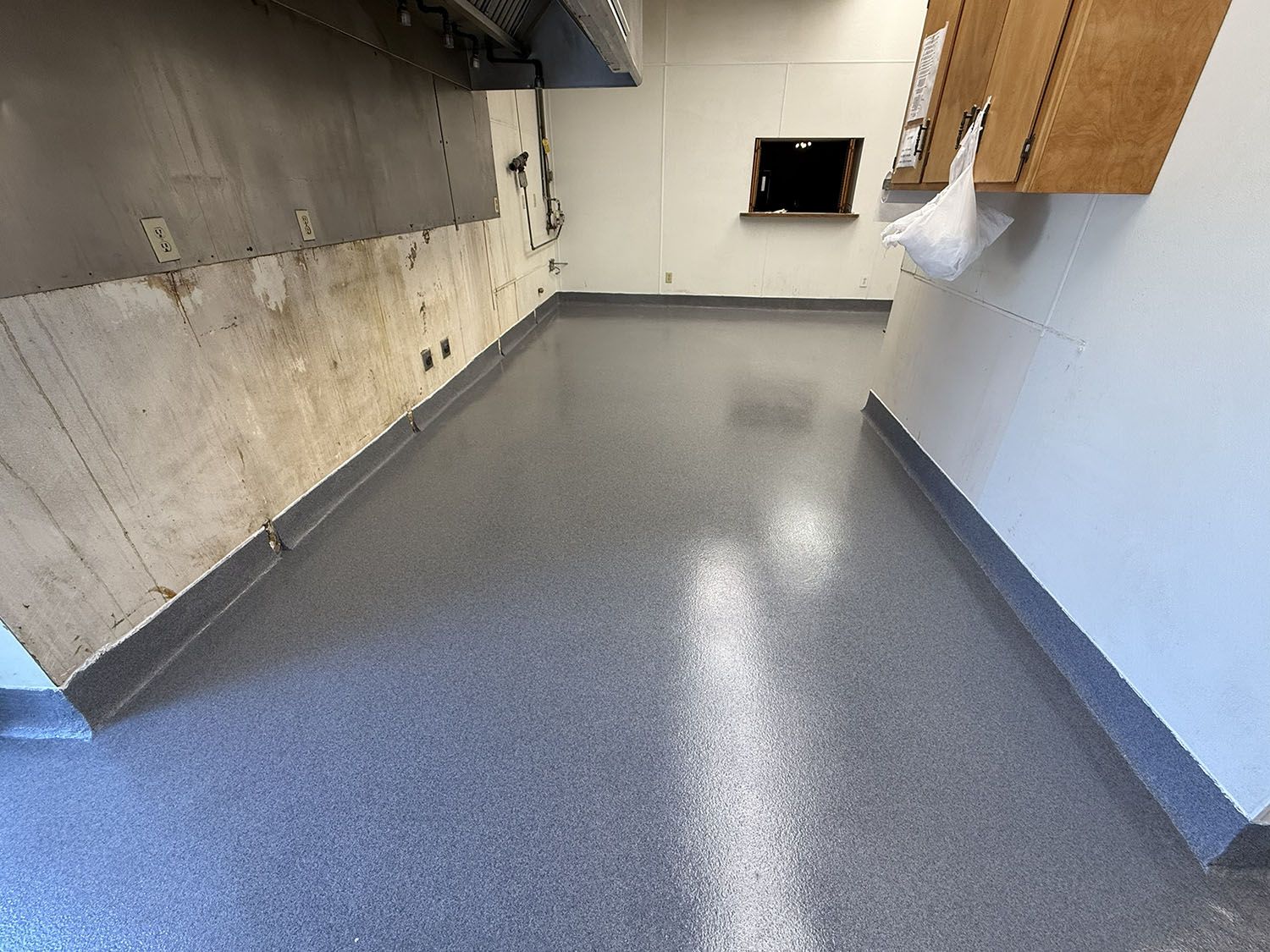 Empty commercial kitchen with gray floor, white walls, and wood cabinets along the right side.