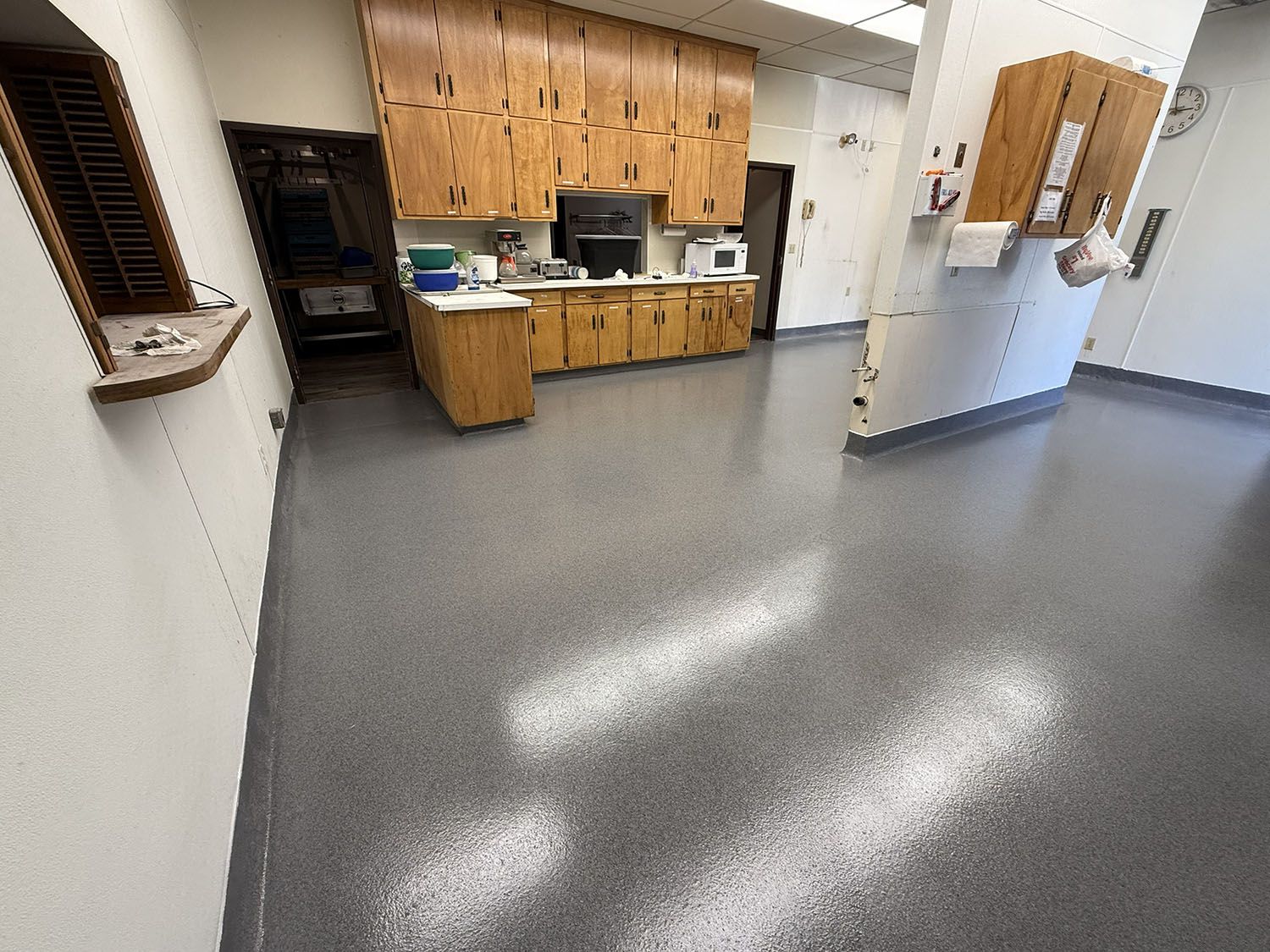 Empty break room with wood cabinets, sink, and gray floor under fluorescent lights.