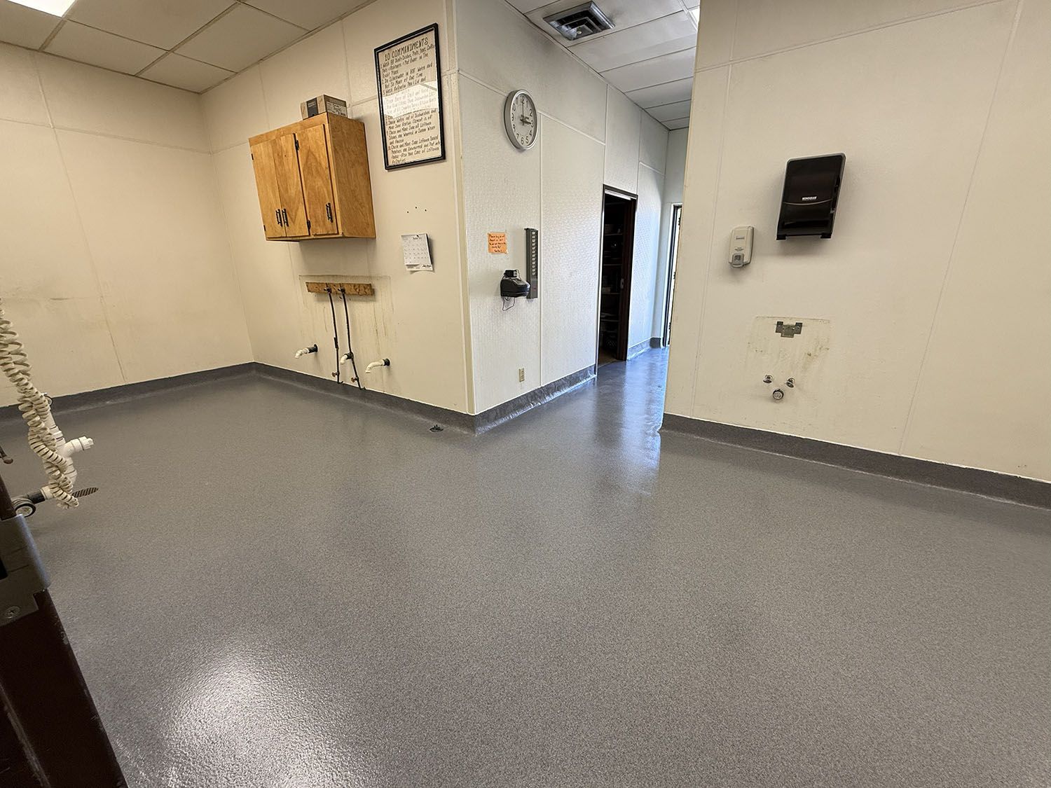 Empty gray-floored hallway with beige walls, wall-mounted cabinets, clock, and paper towel dispenser.