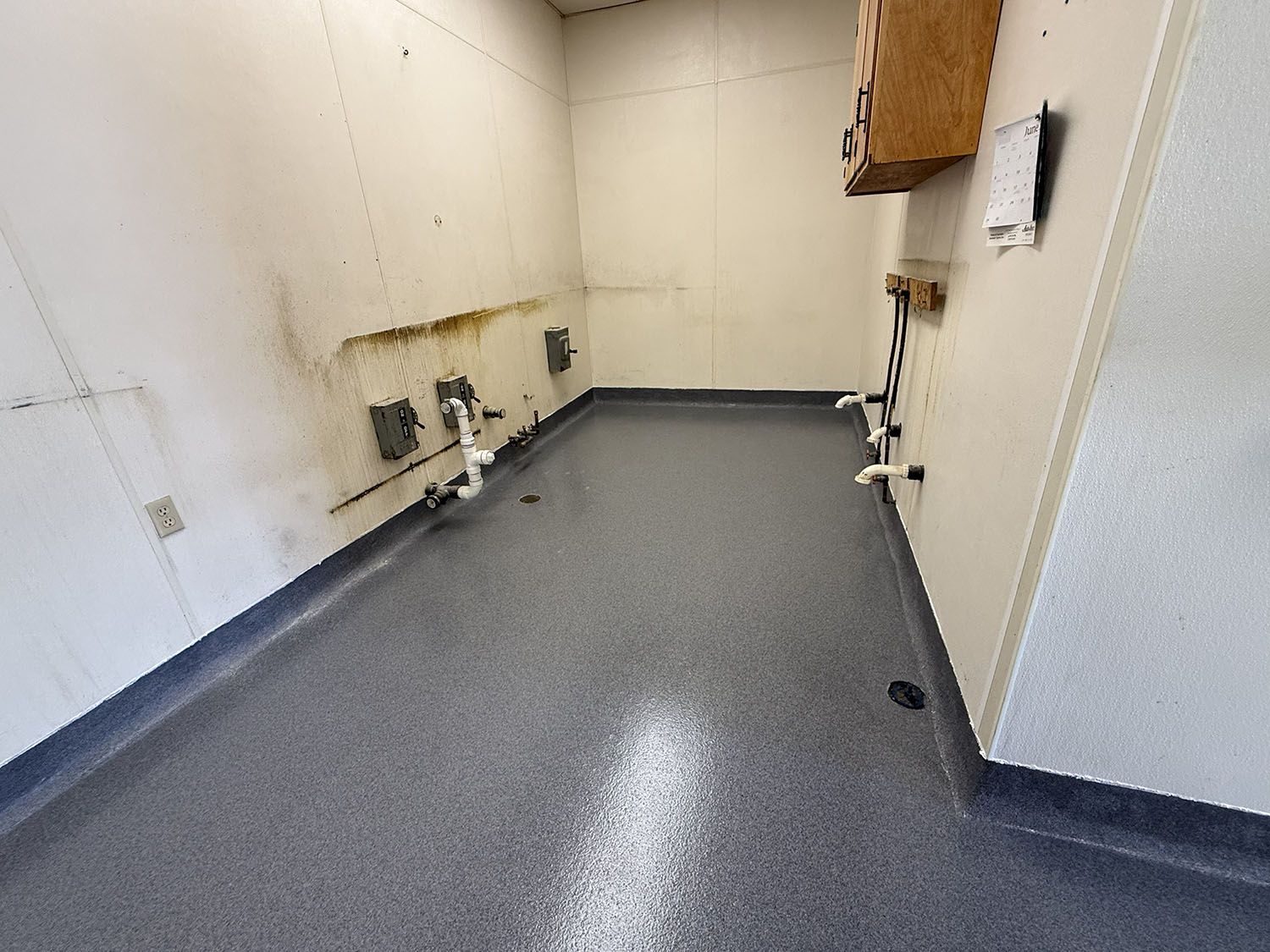 Empty utility room with white walls and gray speckled floor, showing plumbing and electrical fixtures.