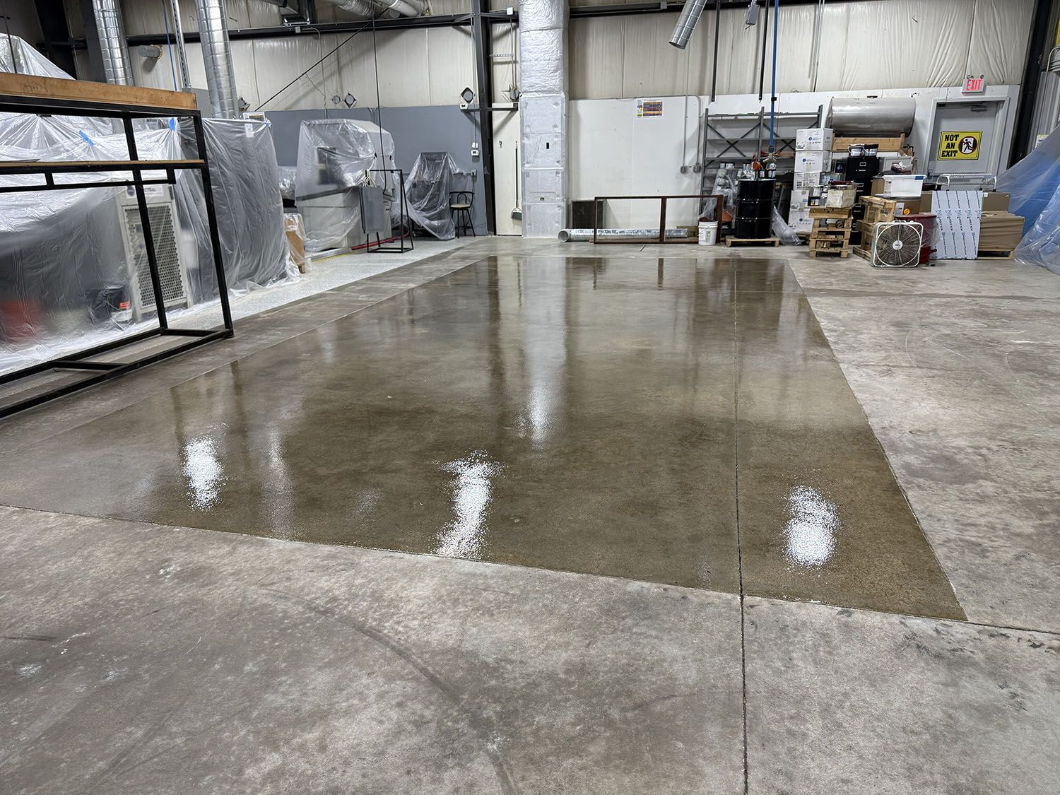 Wet concrete floor in an industrial warehouse, reflecting overhead lights and equipment.