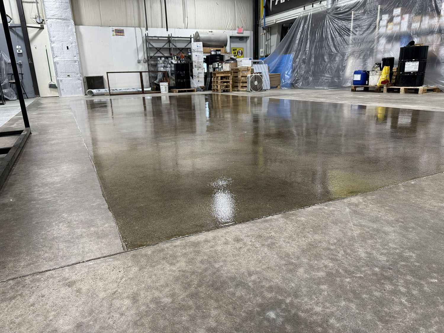Wet concrete floor in an industrial warehouse with equipment and pallets in the background.