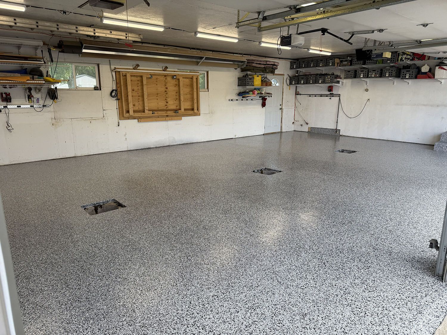 Empty garage with speckled epoxy floor, white walls, and exposed ceiling pipes and lights.