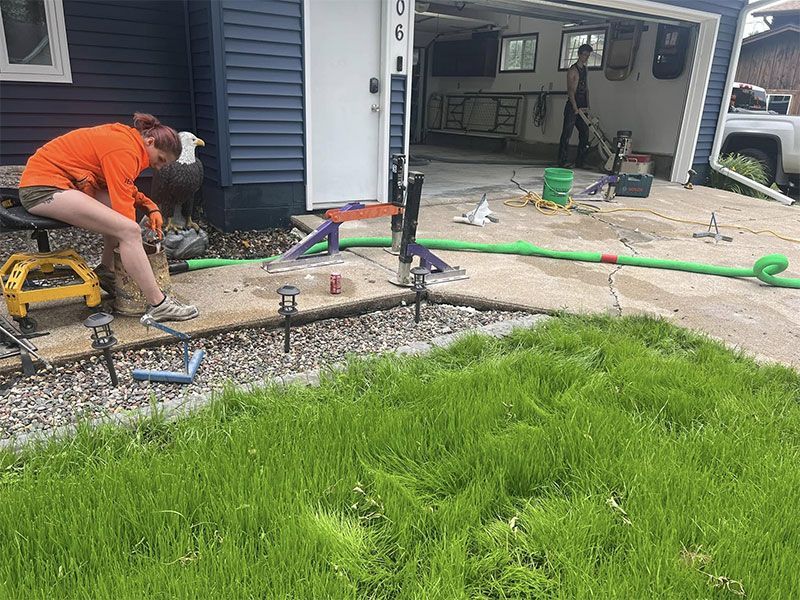 Worker in orange shirt smoothing concrete beside a garage, with tools and green hose nearby