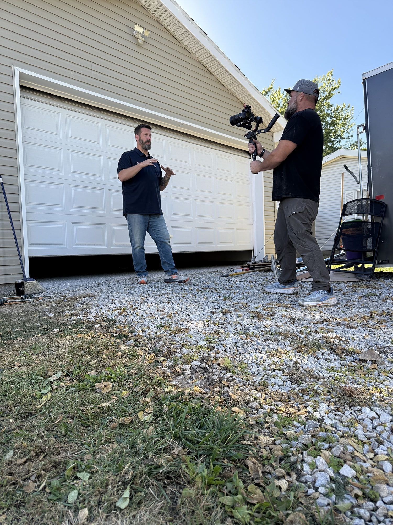 Two men filming outside a garage, one holding a camera on a tripod.