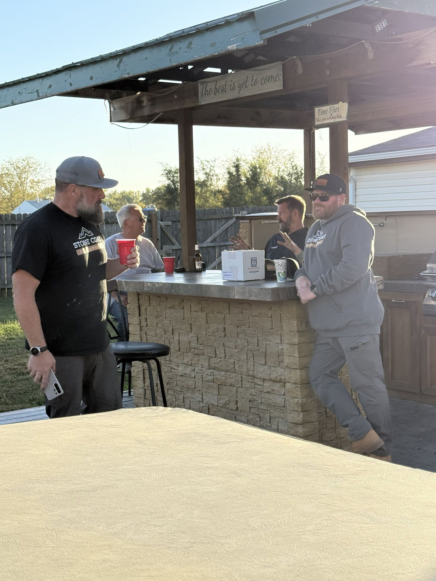 People chatting at an outdoor pavilion bar, with a man in a black shirt and another in gray leaning on the counter.