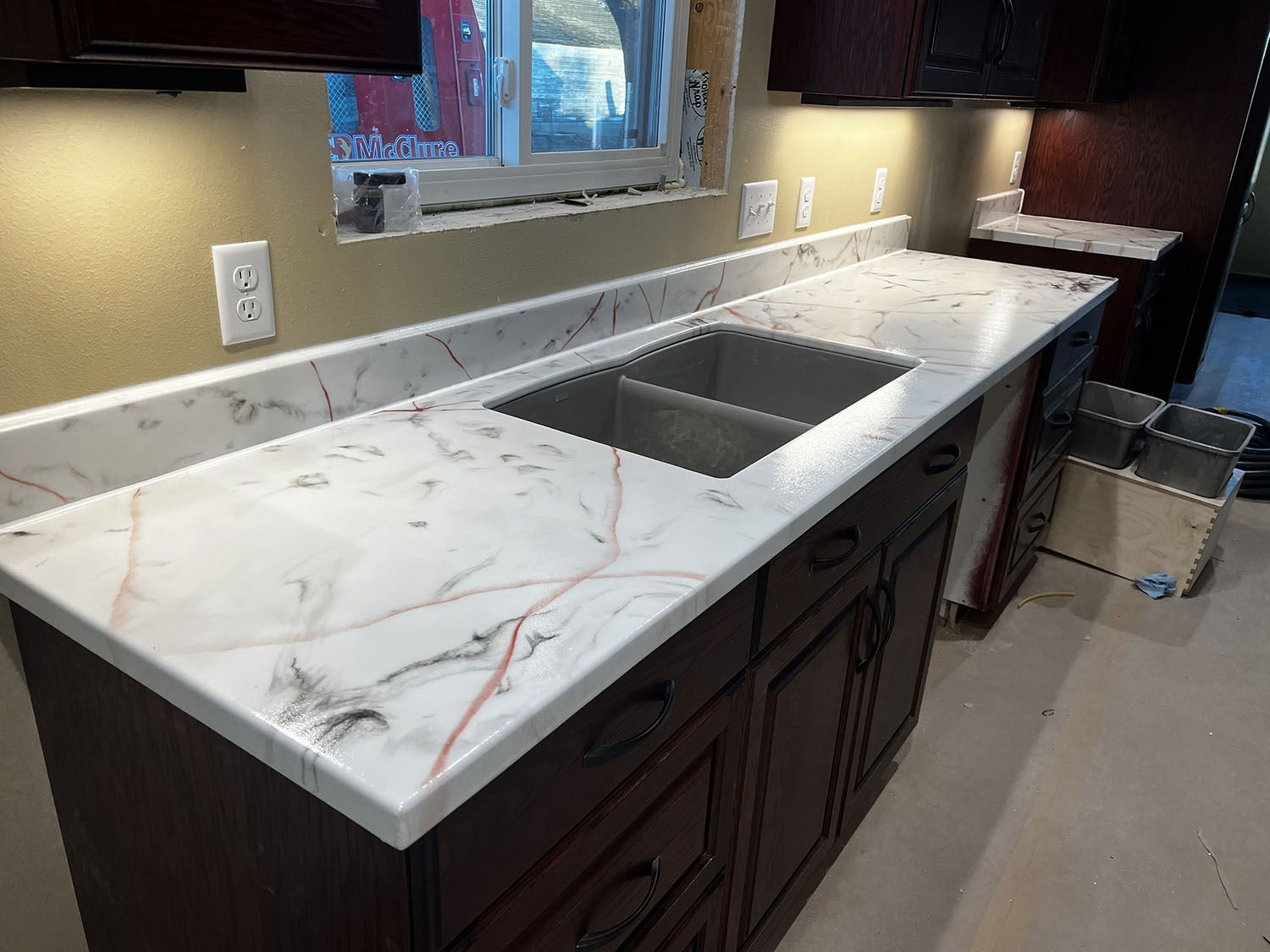Kitchen countertop with white marble look, undermount sink, and dark wood cabinets under a window.