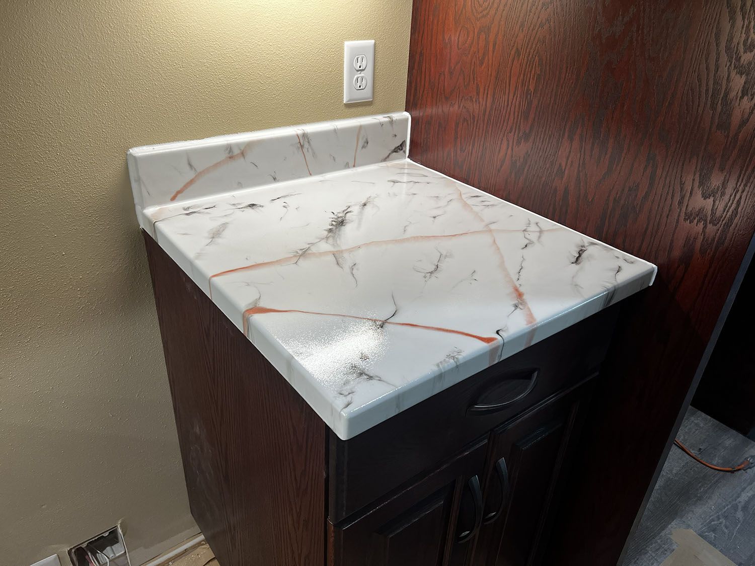 Marble-pattern bathroom vanity countertop with sink cabinet and wall outlet beside a wood-paneled wall.