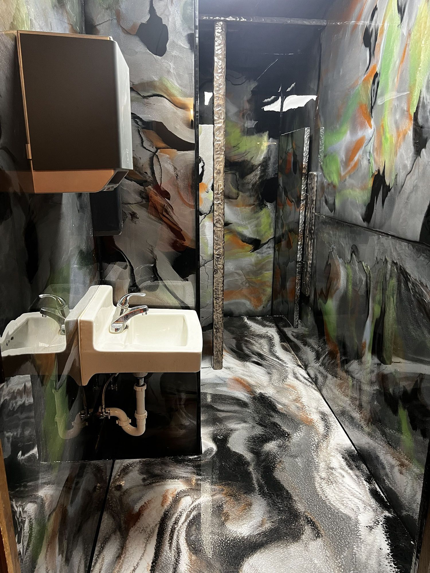 Graffiti-covered narrow bathroom with sink, mirror, and toilet stall in black, white, and orange tones.