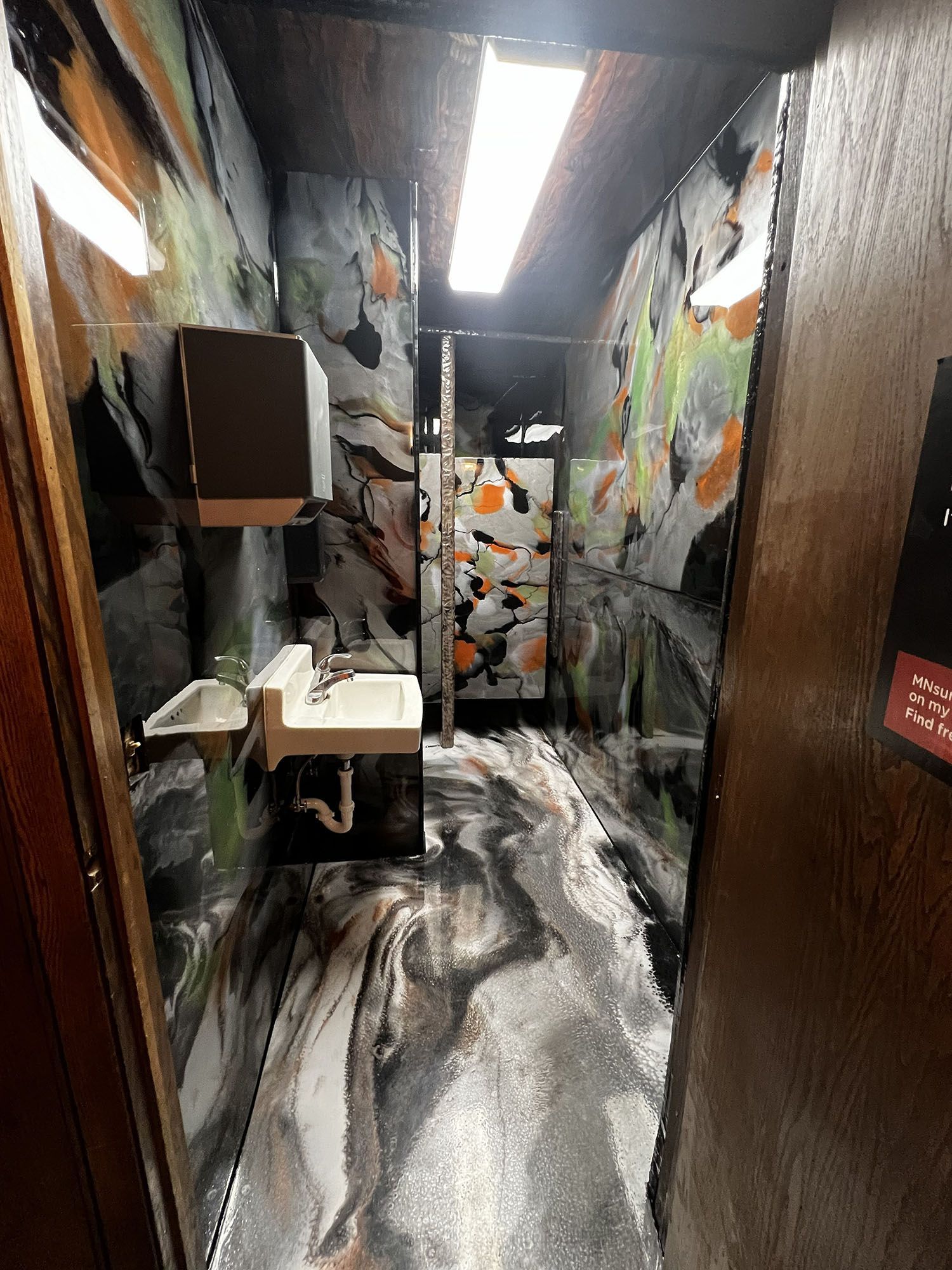 Narrow restroom hallway with graffiti-style walls, sink, mirror, and fluorescent ceiling light.
