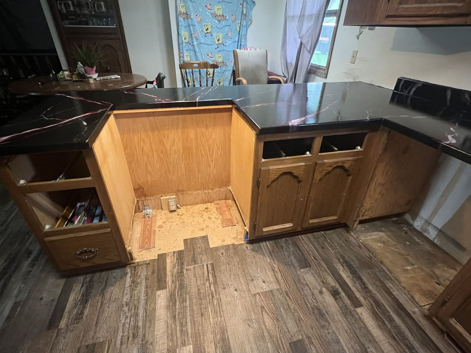 Kitchen with a missing base cabinet section, black countertops, and wood flooring.