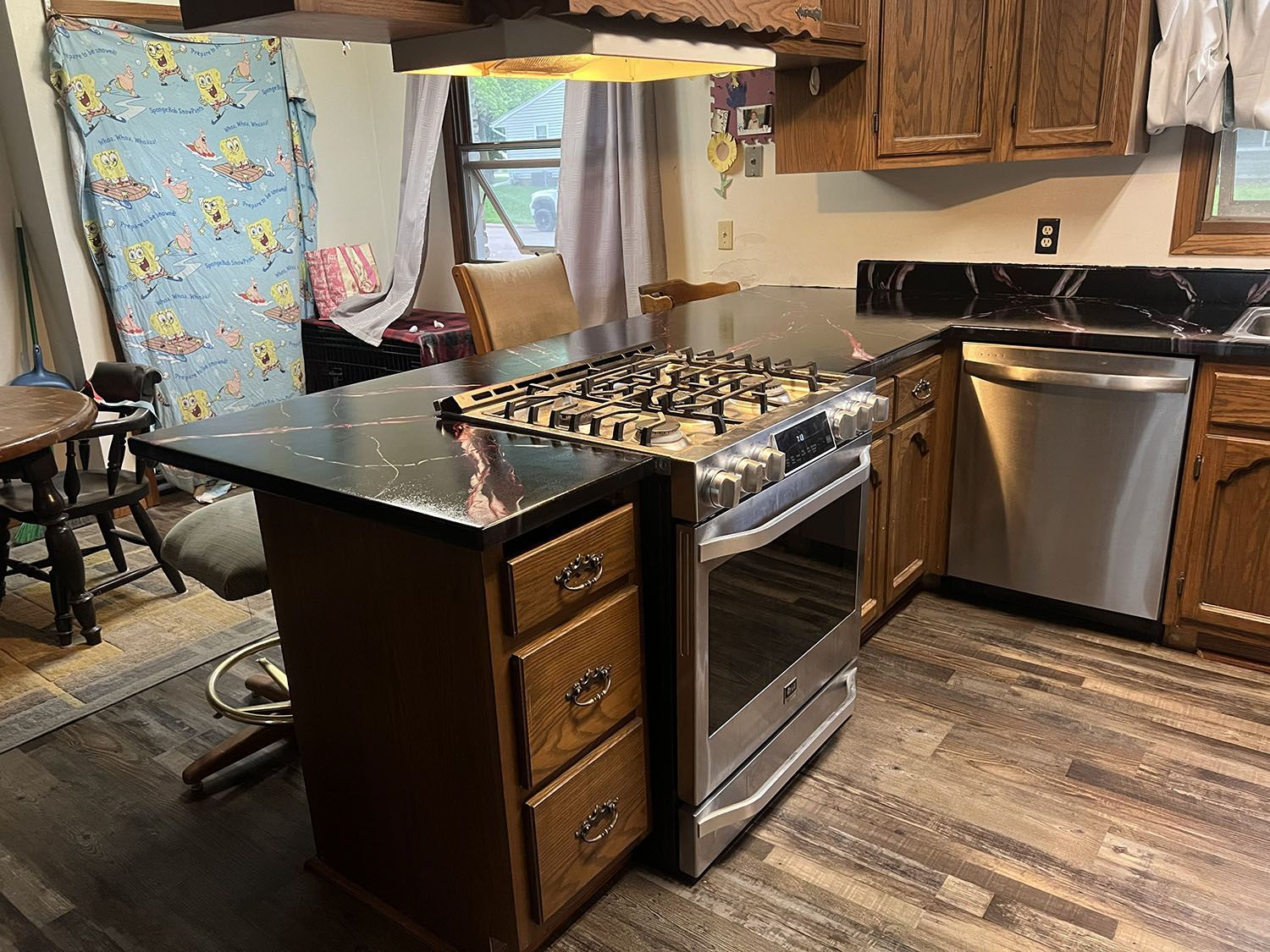 Kitchen with black island, stainless stove and dishwasher, wood cabinets, and floral curtains by the window.