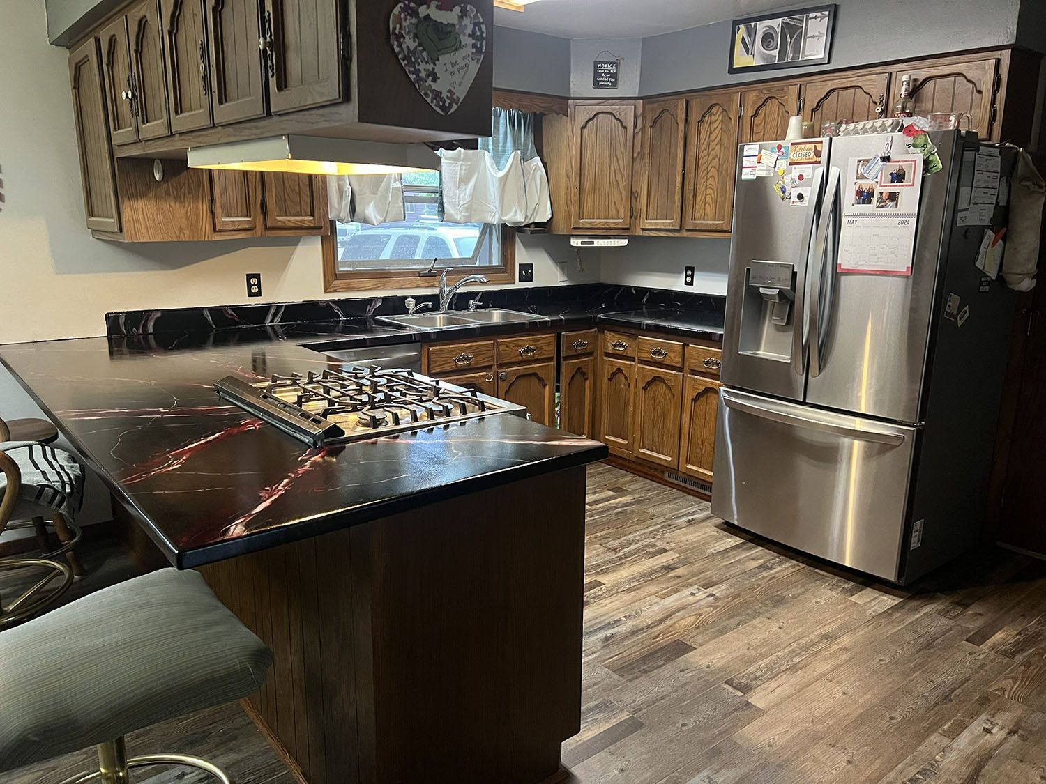 Modern kitchen with black countertops, wooden cabinets, stainless steel refrigerator, and island under pendant lights.