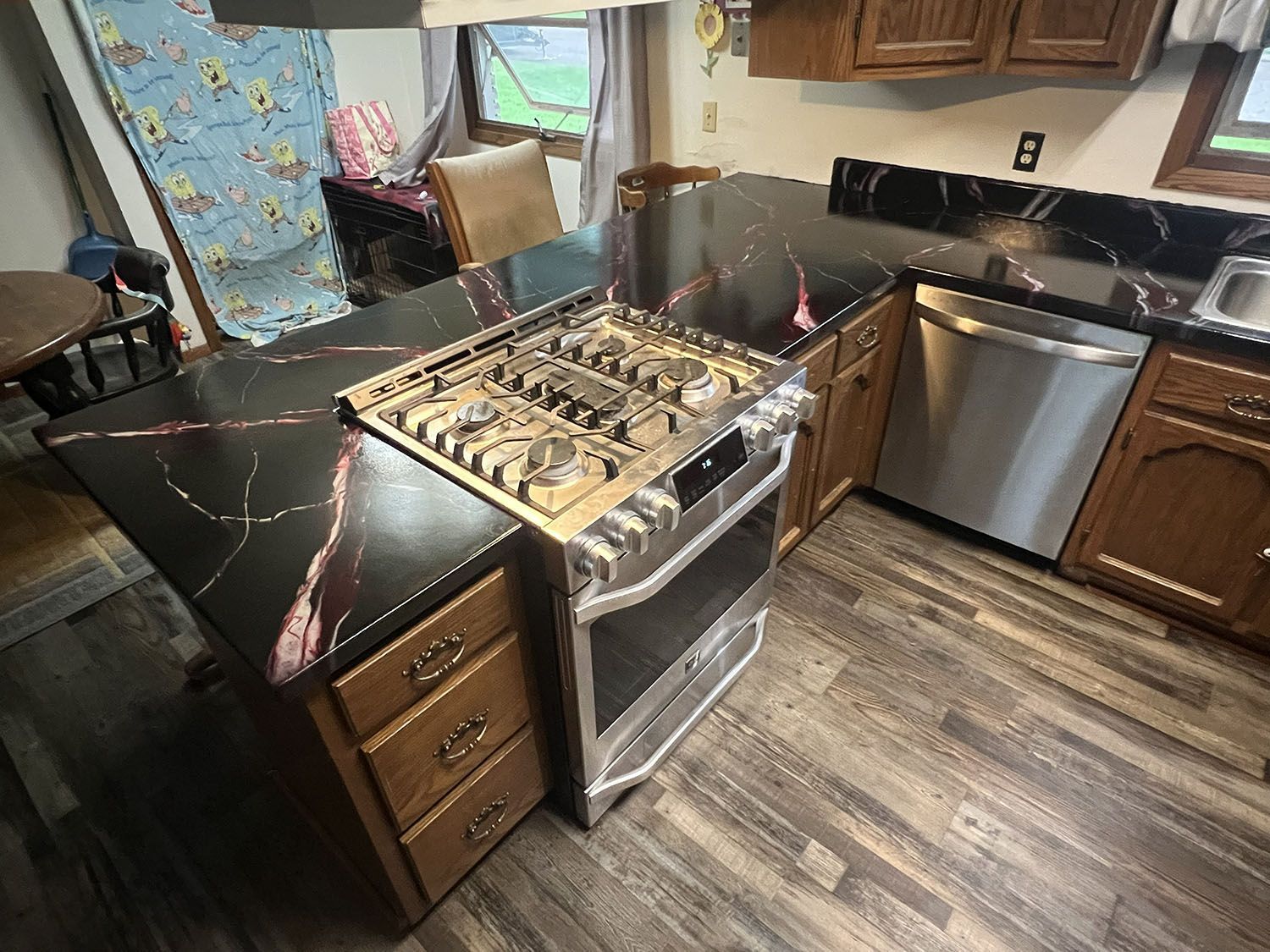 Kitchen with damaged stove, black countertops, wood cabinets, and stainless dishwasher.