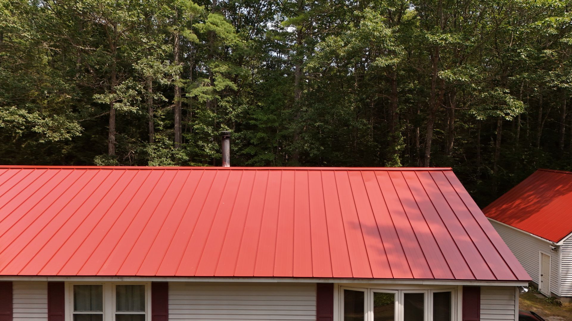 Red metal roof on a white house with a chimney, and a small building; trees in the background.