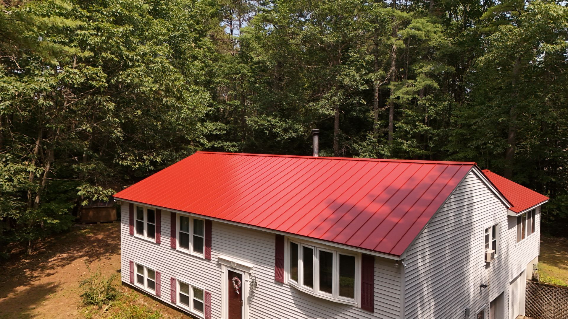 Two-story house with a bright red metal roof and gray siding, surrounded by trees.