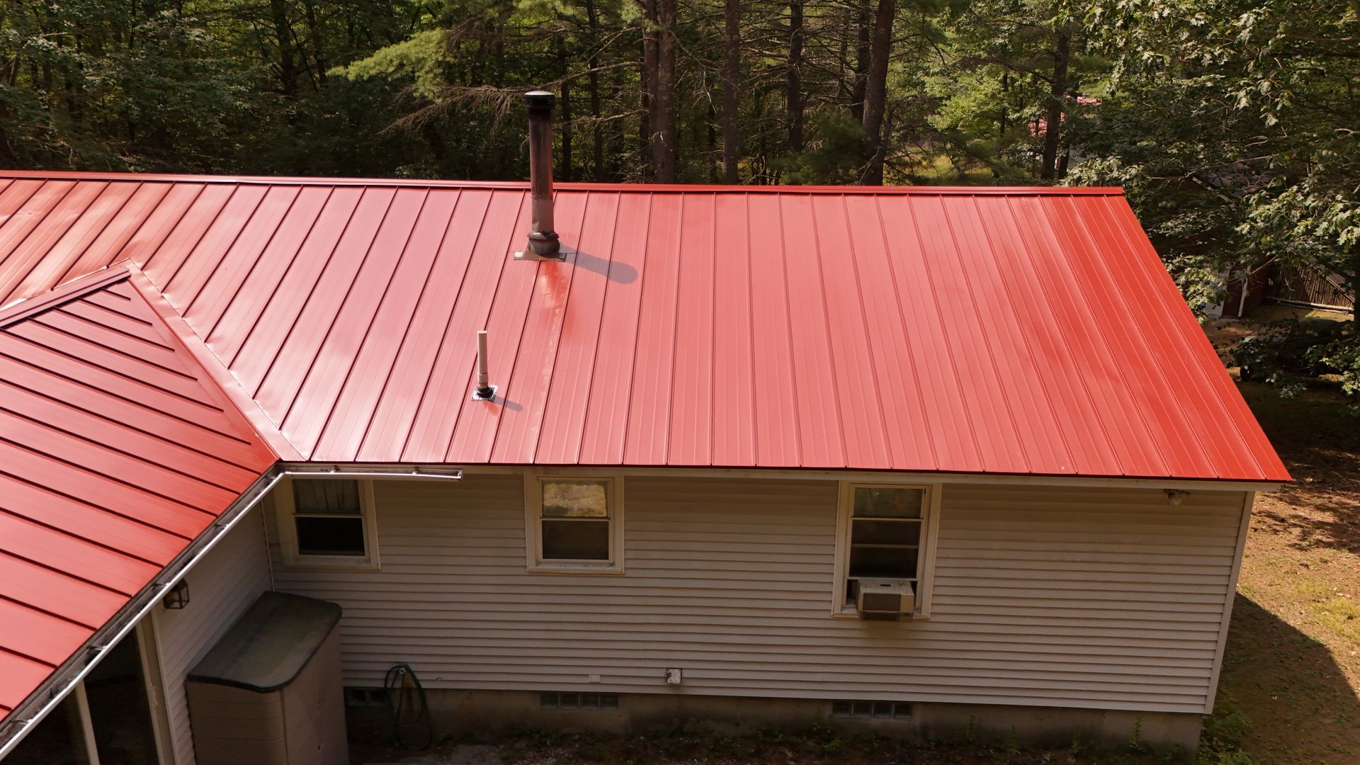 Red metal roof on a white house with a chimney, set in a wooded area.