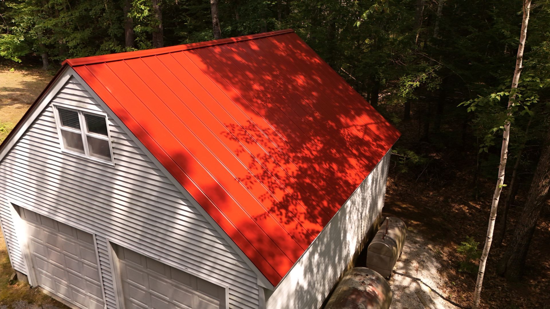 Red-roofed, white-sided shed with two garage doors and a window, surrounded by trees.