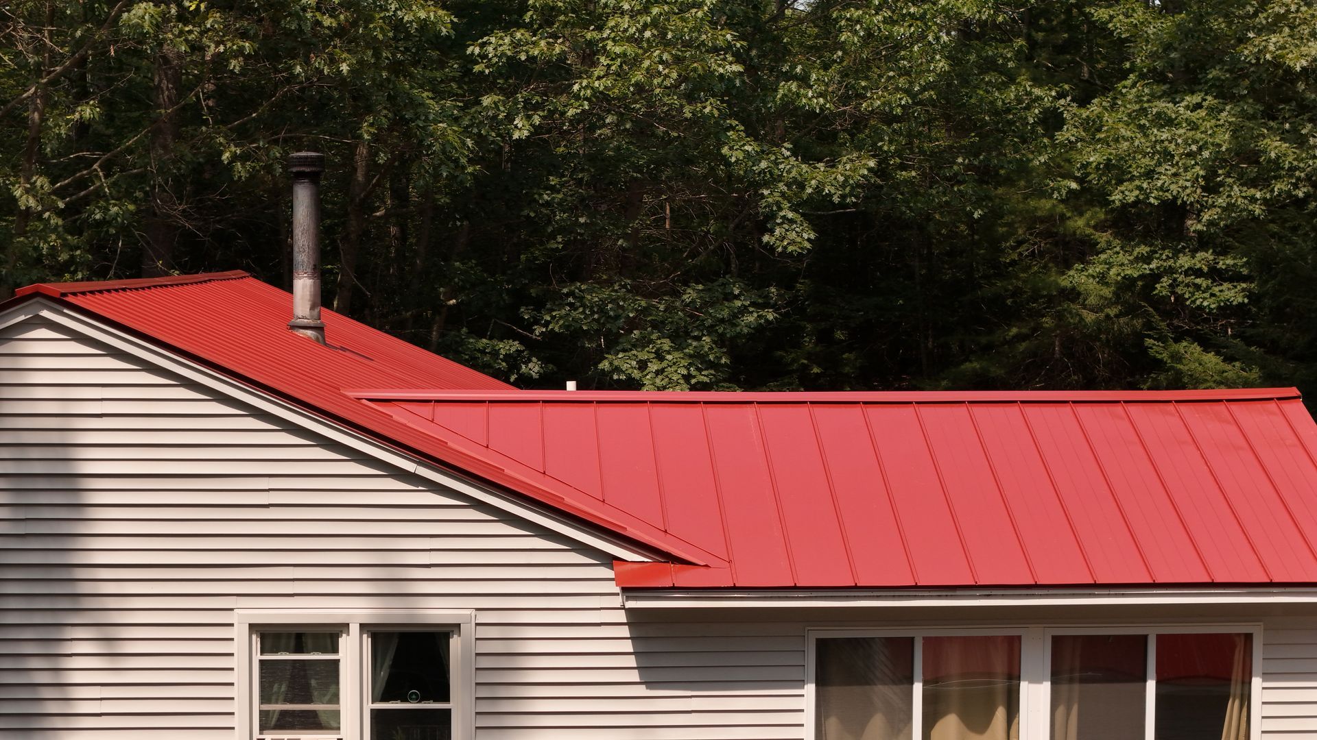 Red metal roof on a gray house with white siding, chimney, and windows; trees in background.