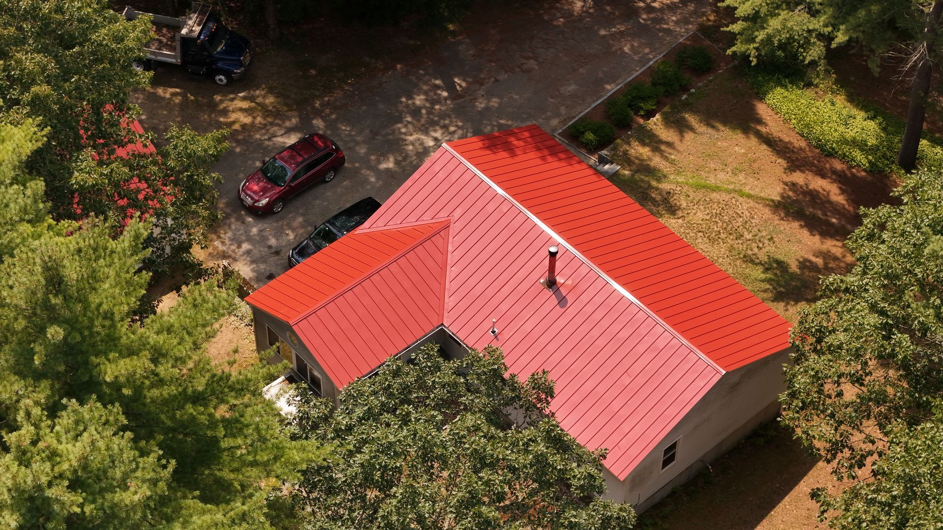 Aerial view of a house with a bright red metal roof surrounded by trees; a red car parked in the driveway.