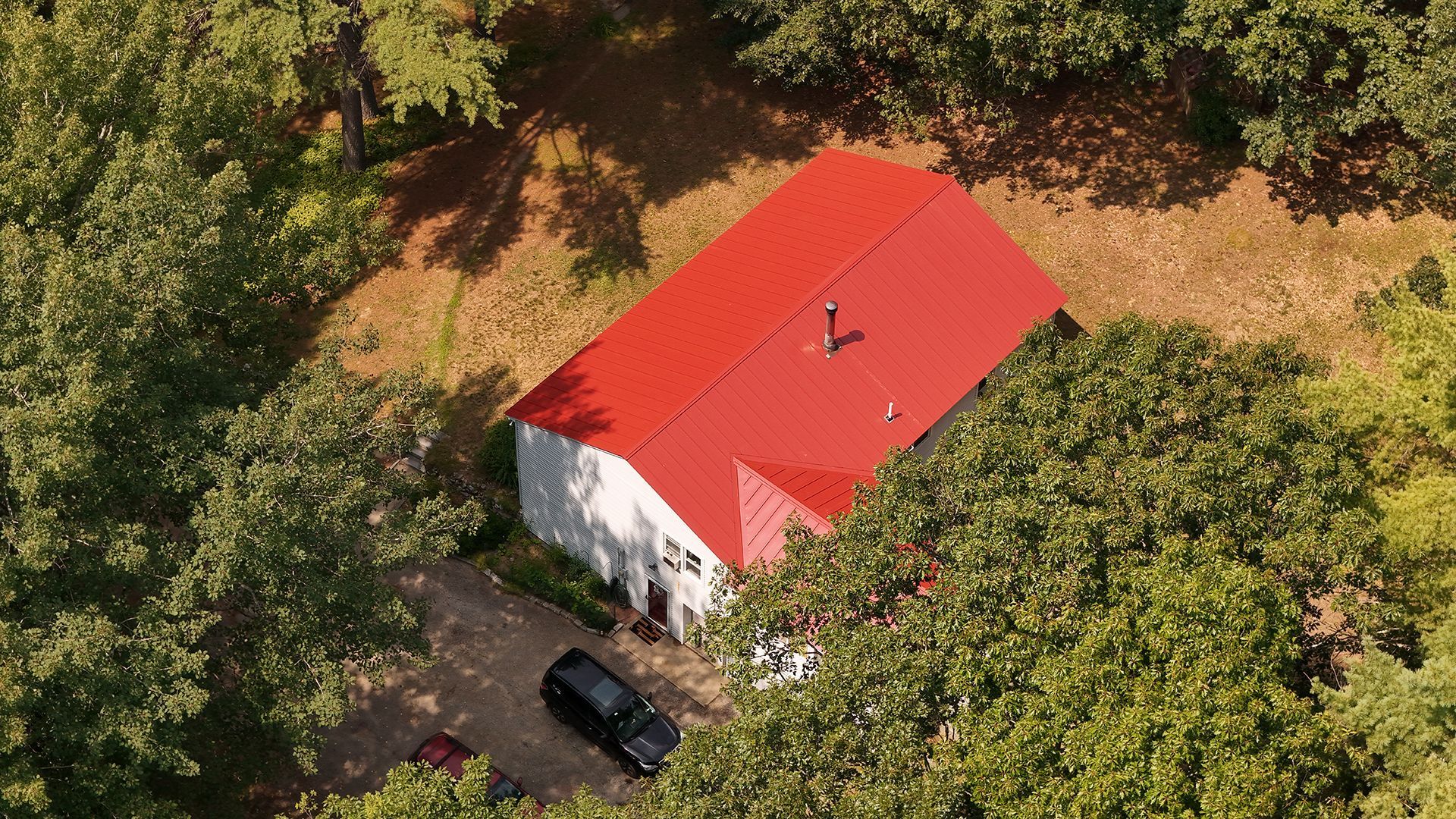 Aerial view of a white house with a bright red roof, surrounded by green trees and a driveway with cars.
