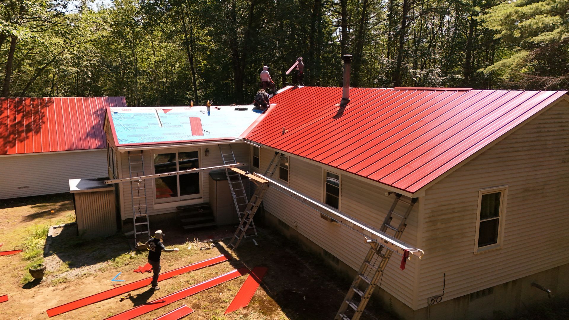 Workers install red metal roofing on a house with ladders and scaffolding.