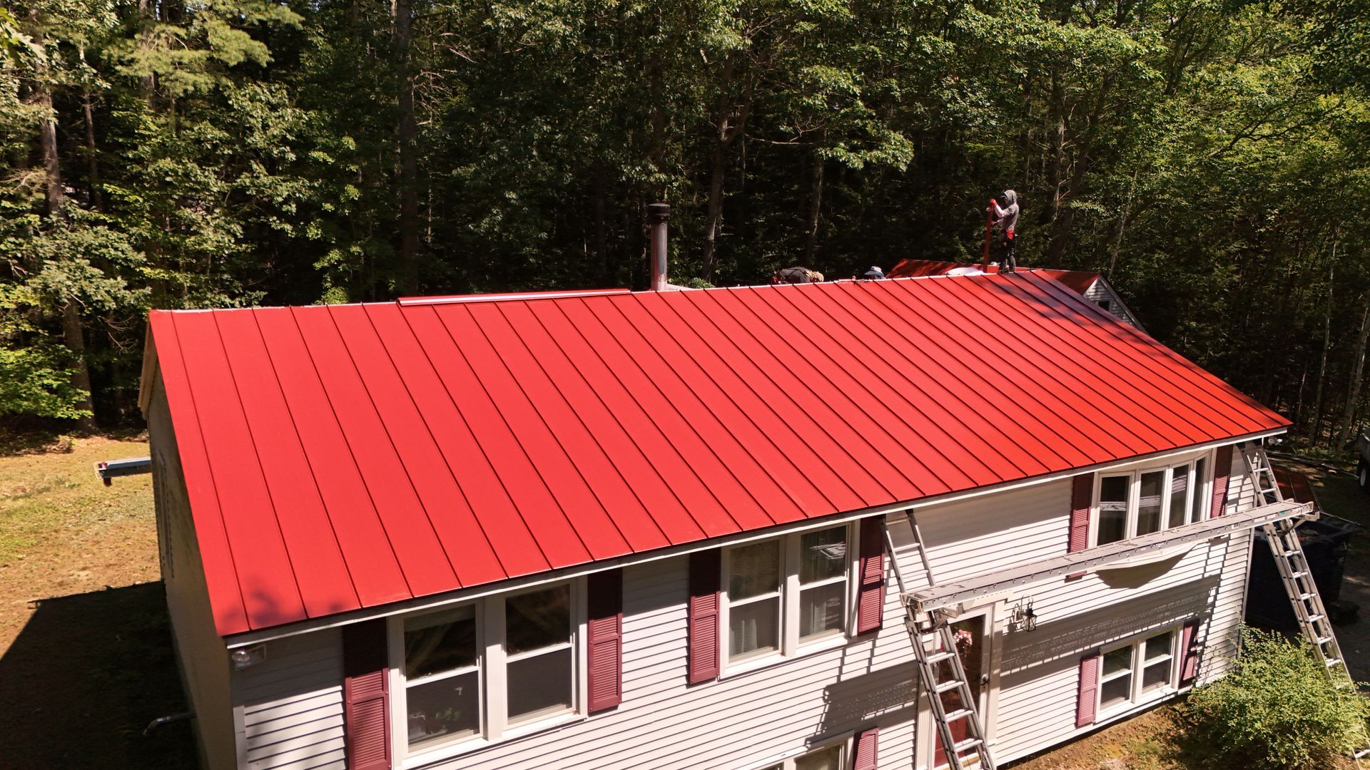 Red metal roof on a two-story house with a person working on it, surrounded by green trees.