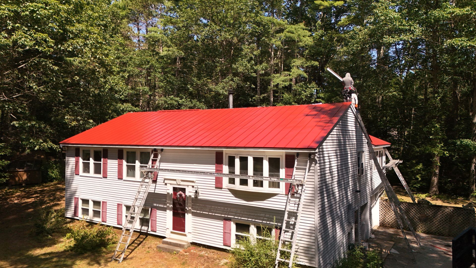 House with red roof, white siding, and red shutters; set in a wooded area with a construction project in progress.