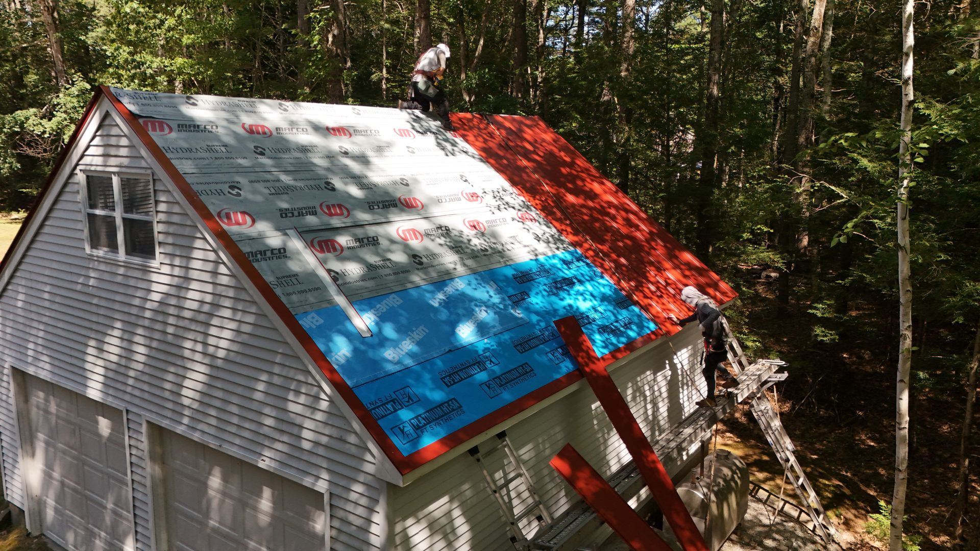 Roofers working on a white building with red and blue underlayment. Forest backdrop.