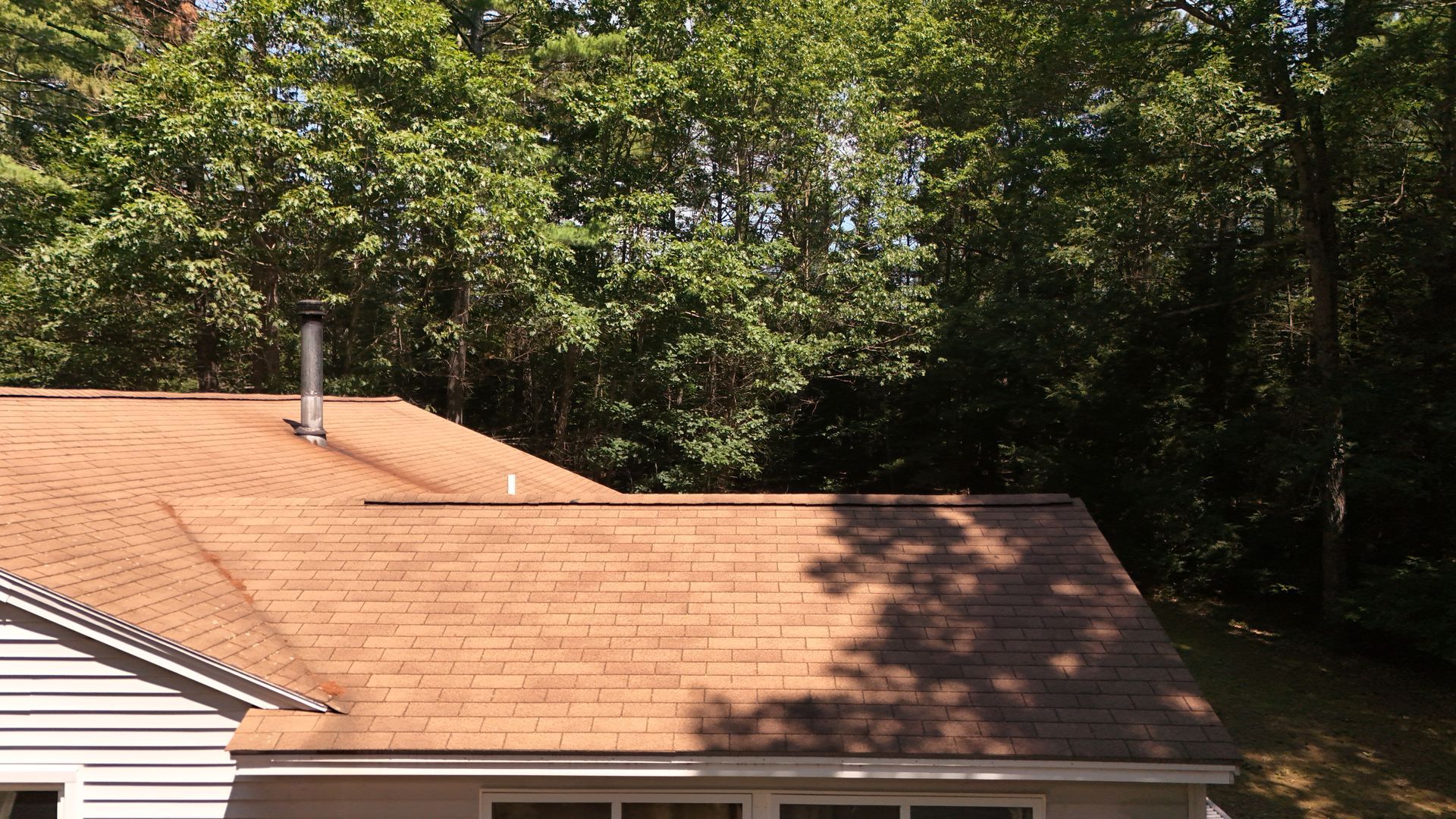 Brown shingled roof with a chimney in front of a backdrop of green trees and a white house.